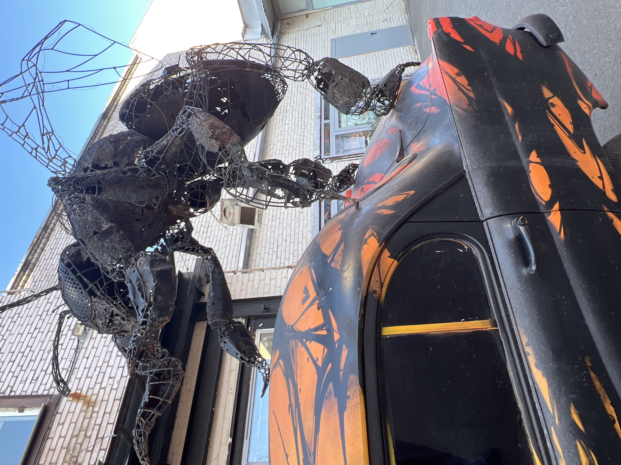 A large, metal sculpture of a bee dominates the left side of the image, hovering near a building and an artistically painted vehicle in what appears to be Saint Petersburg, Russia. The bee, constructed of metal wire and perforated plates, is intricately detailed with its segmented body, wings, and legs clearly visible. It is positioned close to the wall of a brick building with visible windows and an air conditioning unit.
To the right of the bee is a vehicle, painted in black and orange with an abstract design. The vehicle features a rounded door and visible hardware like handles. The angle of the photo is slightly upward, capturing the objects against a bright blue sky.