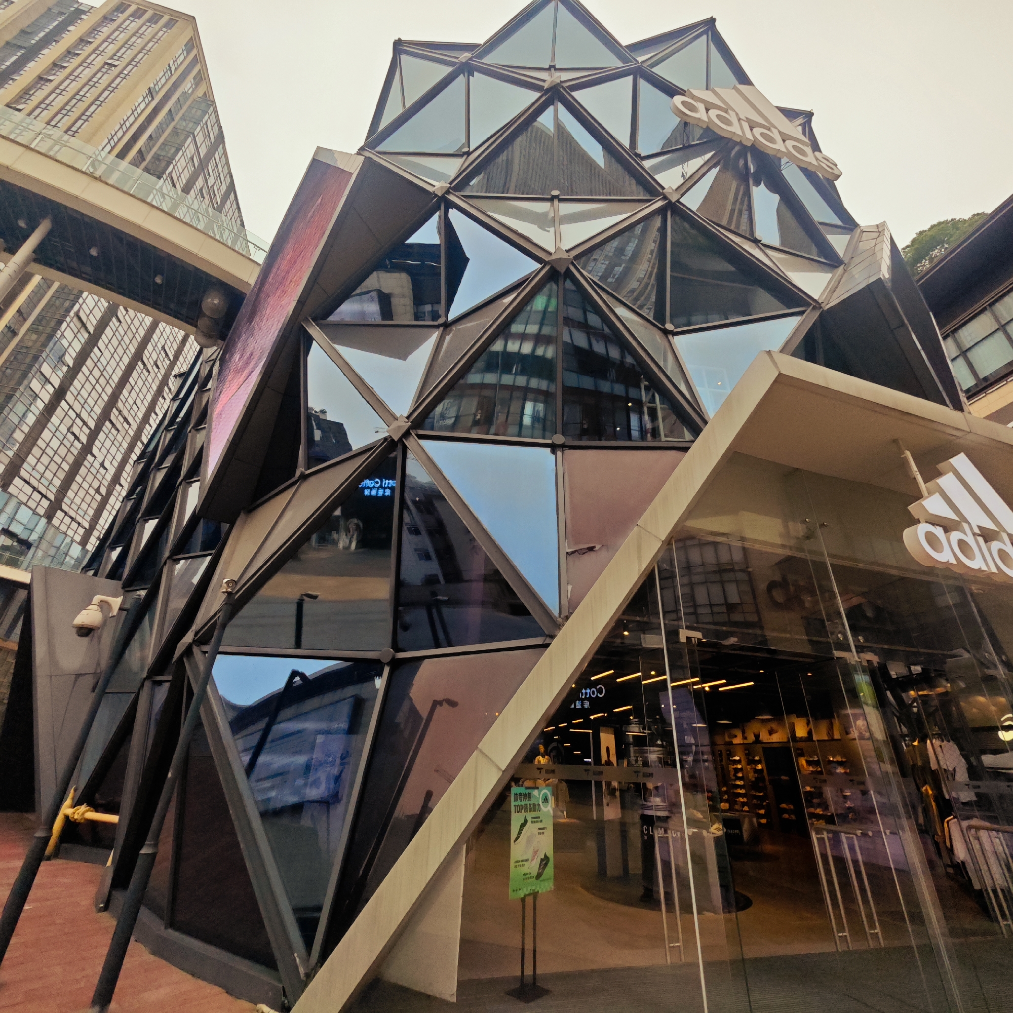 This image presents a modern Adidas store located in an urban setting, likely in Yuzhong District, China. The store features a distinctive, angular facade composed of numerous triangular and polygonal glass panels encased in dark frames, giving it a crystalline or fragmented appearance. A large, white "Adidas" logo is prominently displayed on the upper portion of this geometric structure.
The ground level of the store boasts large glass windows and an entrance, revealing the interior with warm lighting illuminating shelves stocked with shoes and racks displaying clothing. Near the entrance, a vertical green sign stand with Chinese text is visible. The text includes phrases like "酷者冲刺" (Kù zhě chōngcì - Cool runner's sprint) and "TOP者动力" (TOP zhě dònglì - TOP's power/motivation), accompanied by graphics of athletic shoes. Reflections in the glass facade distort views of the surroundings and show reversed text, such as "CLIMATE" and "COTTO," potentially from inside the store or nearby signs.
In the background and to the left, tall, contemporary buildings with many windows rise, reflecting the cloudy sky. An elevated walkway connects parts of these background structures. The lighting suggests an overcast day or perhaps late afternoon, with diffused light creating soft reflections on the glass surfaces. A white security camera is mounted on a wall to the left of the Adidas store. The overall scene captures a dynamic and modern commercial environment, with no discernible individuals in the foreground.
