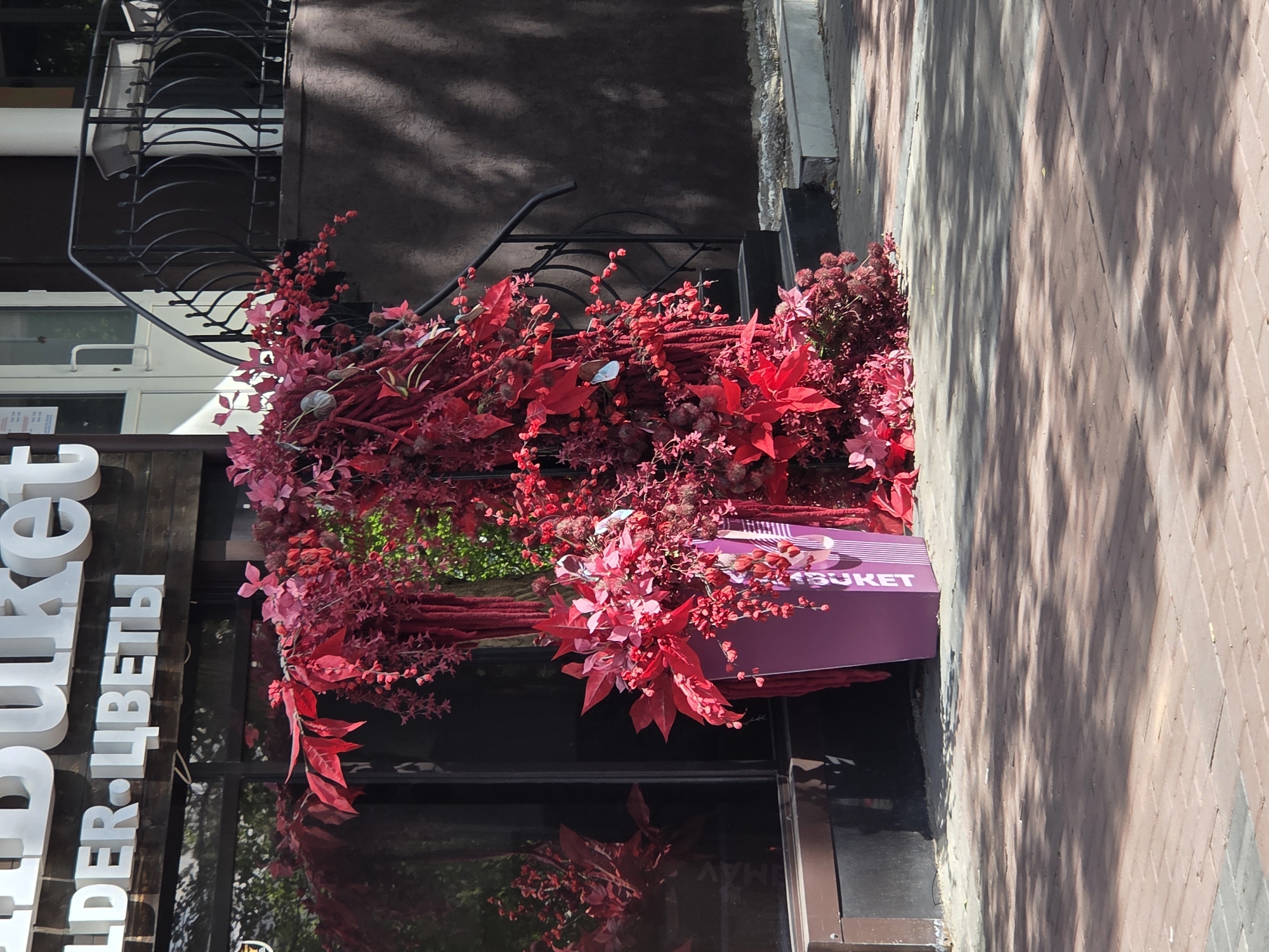 A storefront in Qarağandı city administration, Kazakhstan, is adorned with a large, elaborate floral arrangement of predominantly red and pink foliage and flowers. The arrangement cascades down the side of the building and spills onto the sidewalk. A tall, purple planter with the word "BUKET" visible on its side holds some of the floral display. The storefront has large dark windows reflecting the surrounding environment. A black metal railing with a decorative pattern is visible on a raised section of the building to the right, leading to a doorway. The ground in front of the store is paved with red bricks, and shadows from overhead trees are cast across it. The scene appears to be outdoors on a sunny day.