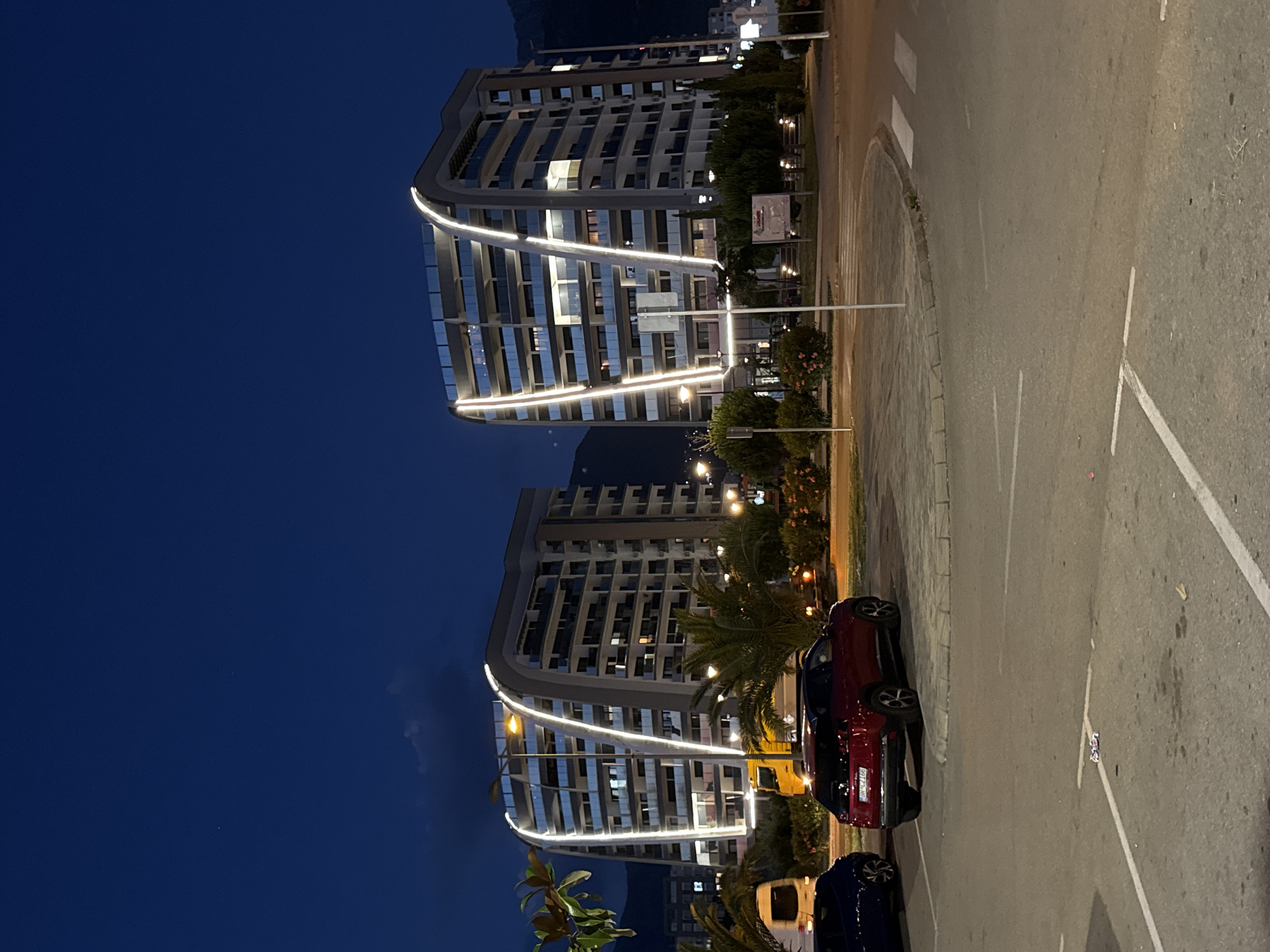 The image depicts two modern apartment buildings with curved facades illuminated by white strip lighting at dusk in the city of Bar, Montenegro. The buildings are tall, multi-story structures with many balconies and windows, some of which are lit from within. In the foreground, a red SUV is parked in a parking lot, and a blue car is partially visible to its left. A yellow truck is also parked behind the red SUV. The surrounding area features palm trees, bushes, and streetlights. The sky is a deep blue, indicating it is evening. The overall scene suggests a residential or mixed-use area in a coastal city.