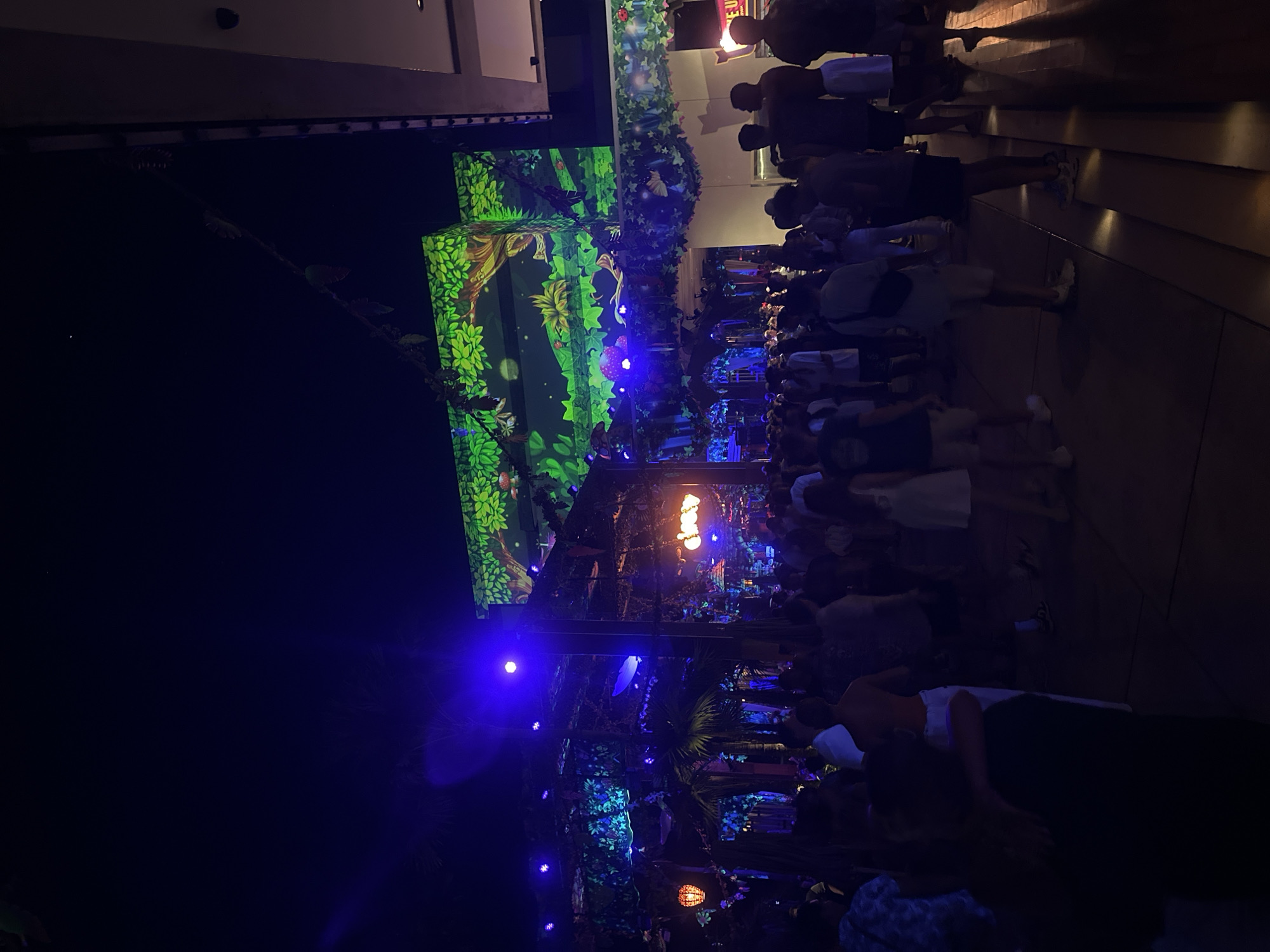 The image depicts a nighttime scene of what appears to be an outdoor event or performance in Kuta Selatan, Indonesia. A crowd of people is gathered, facing a stage or display that features illuminated, cartoonish jungle-themed visuals. The lighting scheme incorporates blue and green hues, with some areas featuring brighter orange or yellow lights. The crowd appears to be diverse in age and attire. The setting has an artificial jungle-like atmosphere.