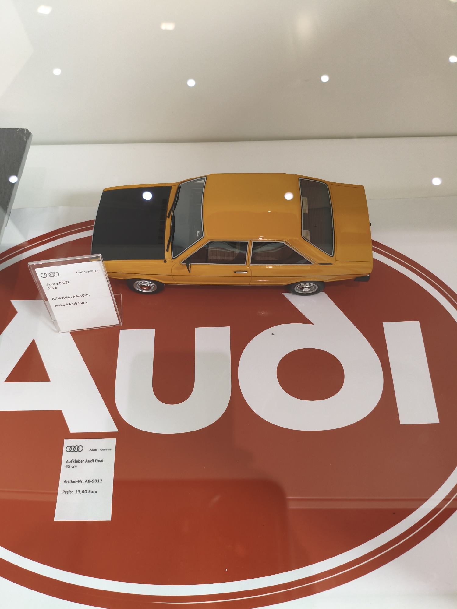 The image shows a yellow Audi 80 GT E model car displayed on a large red and white Audi logo. There are two price tags visible. One tag, in the foreground, lists "Aufkleber Audi Oval 49 cm" with an article number and a price of 13.00 Euro. Another tag, near the car, displays "Audi 80 GTE 1:18" with an article number and a price of 99.00 Euro. The scene appears to be a display within a museum or a showroom in Ingolstadt, Germany, given the prominent Audi branding. The lighting suggests an indoor environment with spotlights. No people are visible in the image.