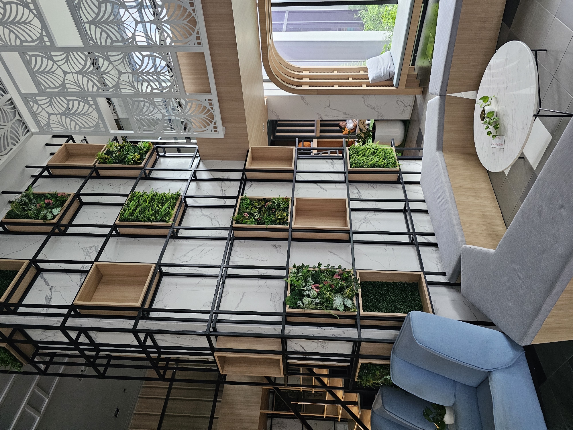 This is a photograph of a modern interior space, likely a cafe or lounge, with an emphasis on natural elements and contemporary design. The scene is dominated by a large, wall-mounted shelving unit filled with a mix of empty wooden boxes and planters containing artificial greenery. The planters are arranged in a grid-like pattern against a backdrop of marble-patterned tiles.
In the upper portion of the image, a decorative white metal screen with a leaf motif is visible, adding an architectural detail. To the right, a circular white marble table with a small potted plant sits near a light blue armchair and a gray sofa. The seating appears to be arranged to face outward or towards a large window, suggesting a place for relaxation and conversation.
The lighting suggests it is daytime, with natural light entering from the top and right side of the frame. The overall impression is one of a clean, stylish, and inviting atmosphere, possibly located in a commercial area of Bangkok, Thailand, given the context. There are no people clearly visible in the frame, and no discernible text is present. The orientation of the photo suggests it was taken from a slightly elevated angle, looking down and across the space.