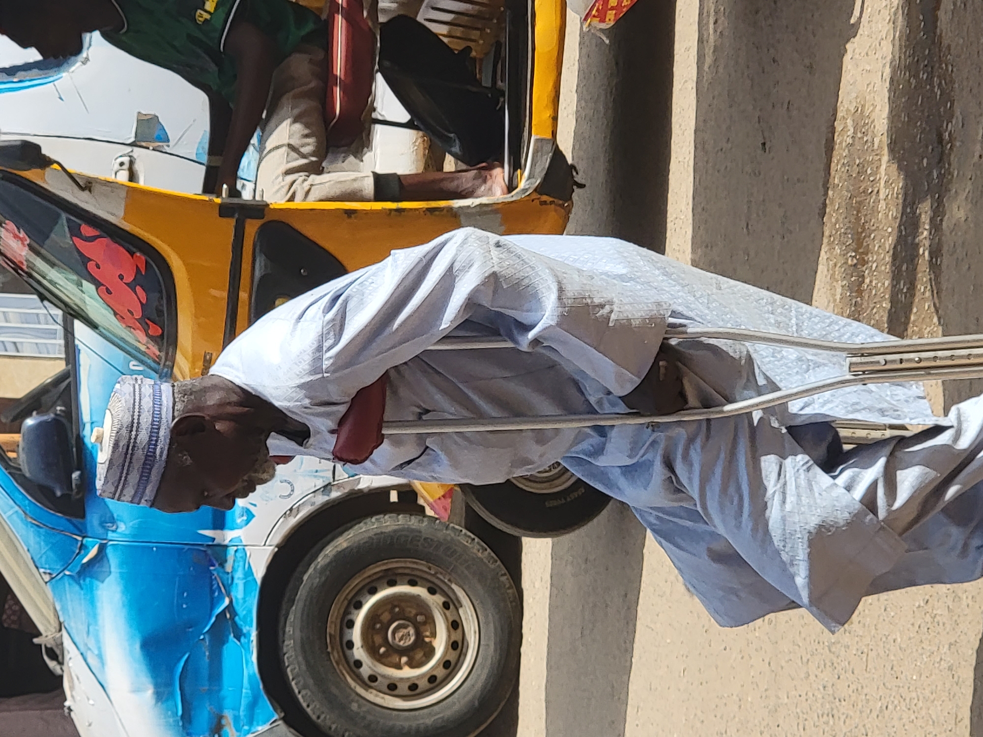 An outdoor scene in Maiburiji, Nigeria, depicts a man standing with crutches next to an auto-rickshaw, commonly known as a "Keke Napep." The image is oriented vertically but depicts a man upright, wearing a light blue/grey traditional garment and a matching cap, leaning on two metal crutches. His head is visible, showing a dark complexion and some facial hair.
Behind him, to the left, is a blue and yellow auto-rickshaw. The blue section of the vehicle, particularly the lower part of the side panel, shows wear and some damage, including peeling paint and dents. Red, flame-like decals adorn the yellow upper section near the front window. Inside the auto-rickshaw, another person is partially visible, wearing a green shirt. Their arm and leg can be seen, and they appear to be seated.
The scene is brightly lit by what appears to be strong sunlight, suggesting it is daytime. The ground and a wall in the background are light brown/beige, characteristic of an arid or dusty environment. One of the vehicle's tires is visible, with "Bridgestone" faintly legible on its sidewall. The overall impression is a common street or roadside scene in a Nigerian town, showing daily life and modes of transport.