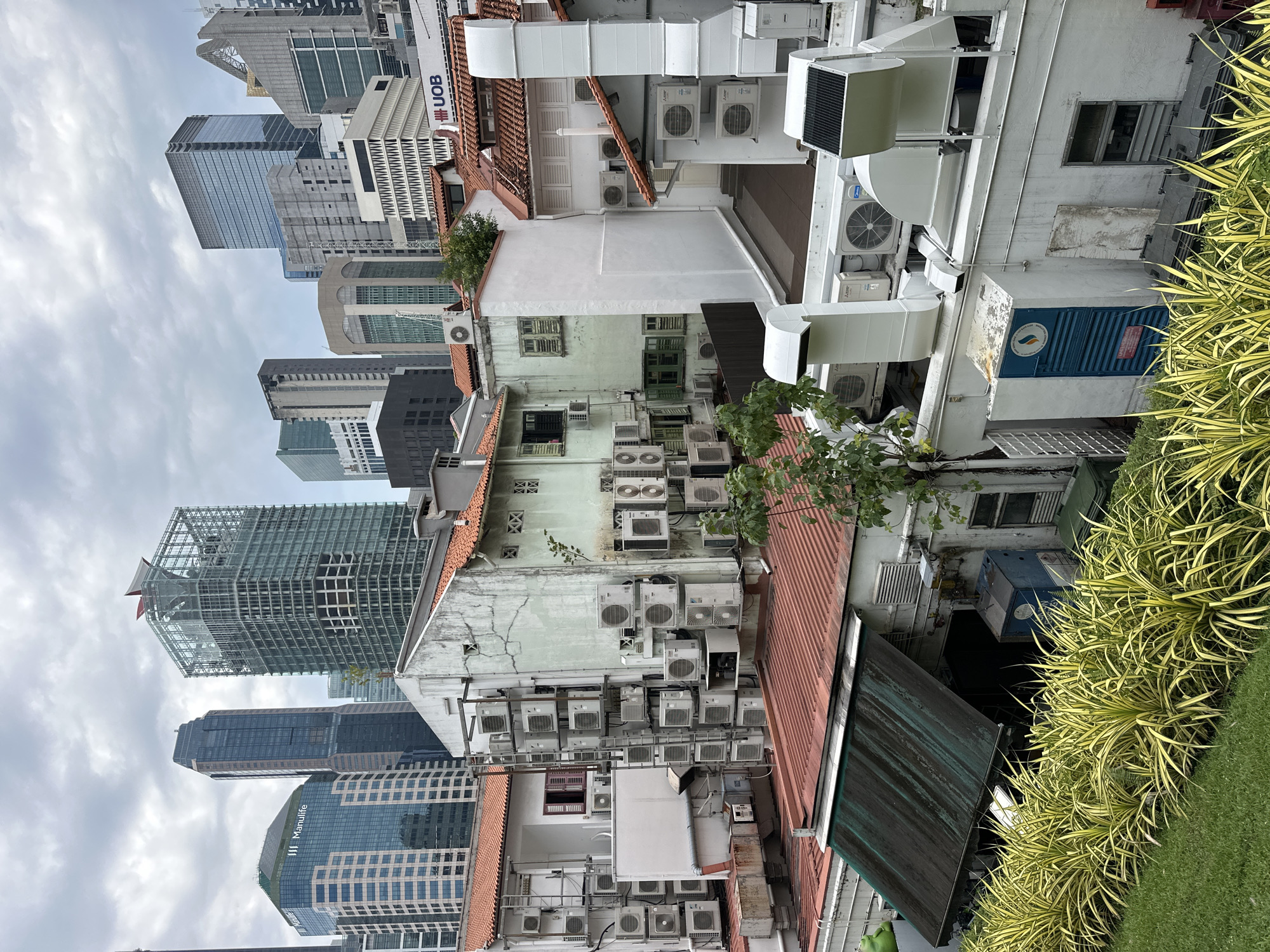 The image depicts an urban scene in Singapore, characterized by a juxtaposition of modern skyscrapers and older, low-rise buildings densely packed with air conditioning units. In the background, several tall, contemporary office buildings dominate the skyline under a cloudy sky. One prominent building on the left features the "Manulife" logo. Closer to the foreground, a series of aging, multi-story buildings with terracotta tiled roofs are covered with numerous external air conditioning units, mounted on walls and scaffolding. These units are of various sizes and brands. The buildings have aged exteriors, with visible wear and tear on their walls. The foreground features lush green grass and ornamental plants with yellow and green foliage. There are no people visible in the image. The environment appears to be a densely populated urban area, likely a commercial or mixed-use district, with a heavy reliance on air conditioning systems.