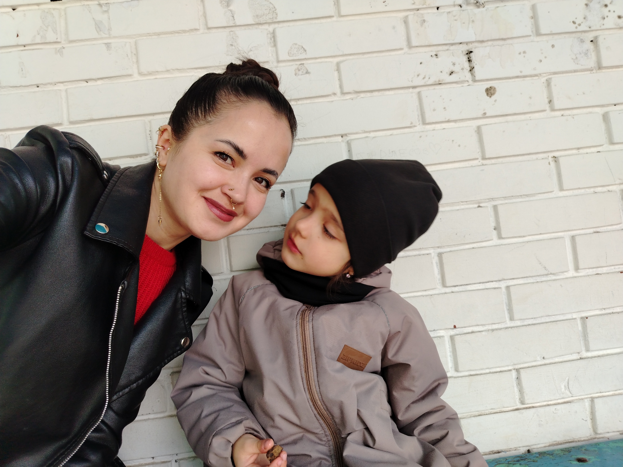 A woman and a young child are posing for a photo in front of a white brick wall. The woman, on the left, is wearing a black leather jacket over a red shirt. She has her dark hair pulled back in a bun and is wearing jewelry in her ears and nose. The child, on the right, is wearing a grey hooded jacket and a black beanie. They have a black scarf around their neck. The child is looking towards the woman. The photo was taken in Noviy, Russia.