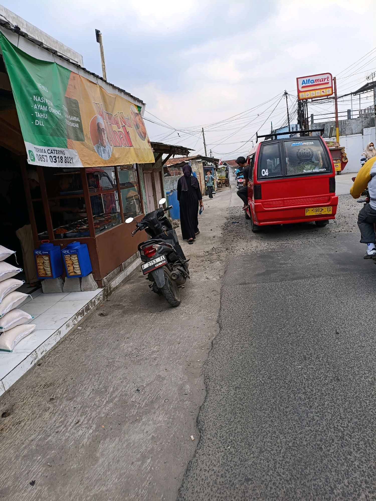 The image depicts a street scene in Sukaraja, Indonesia, under an overcast sky. On the left, a food stall with a green and yellow banner advertises catering services and menu items like "Nasi Putih," "Ayam Goreng," and "Tahu." A phone number, 0857 0318 8021, is prominently displayed. Next to the stall, two blue containers are stacked, and several white sacks of rice are arranged neatly. A black motorcycle with license plate "F 2753 TX" is parked on the right side of the stall.
In the center of the street, a red van with the number "04" on its rear window and the text "Pesona dedamuda" is parked. A person is seated inside the van. Further down the street, a woman in a black hijab walks away from the camera. To the right of the van, a person on a motorcycle, wearing a yellow jacket and grey pants, is also present.
In the background, an Alfamart sign is visible, indicating a commercial establishment. Power lines crisscross the sky, and various buildings line the street, suggesting a typical urban or semi-urban environment. The road surface appears to be asphalt with some visible patches and wear.