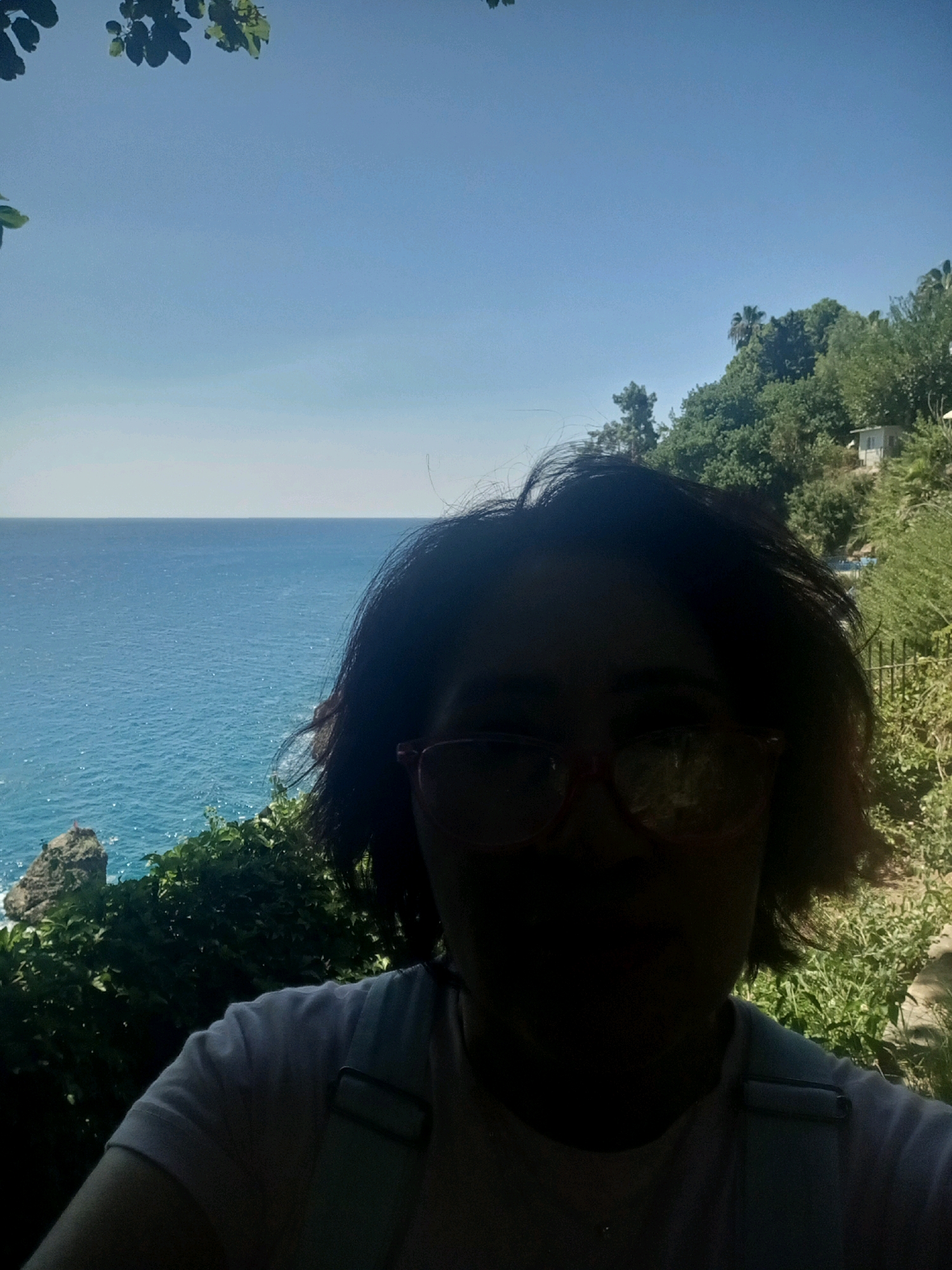 A selfie is taken by a person in the foreground, with their face largely obscured by shadow. They are wearing glasses with red frames and a light-colored t-shirt with suspenders. Behind them, a bright blue sea stretches to the horizon under a clear sky. Lush green vegetation and trees cover a hillside to the right, with a small white building visible among the trees. A rocky outcrop is visible in the sea to the left. The scene is consistent with a location along the coastline of Antalya, Turkey.