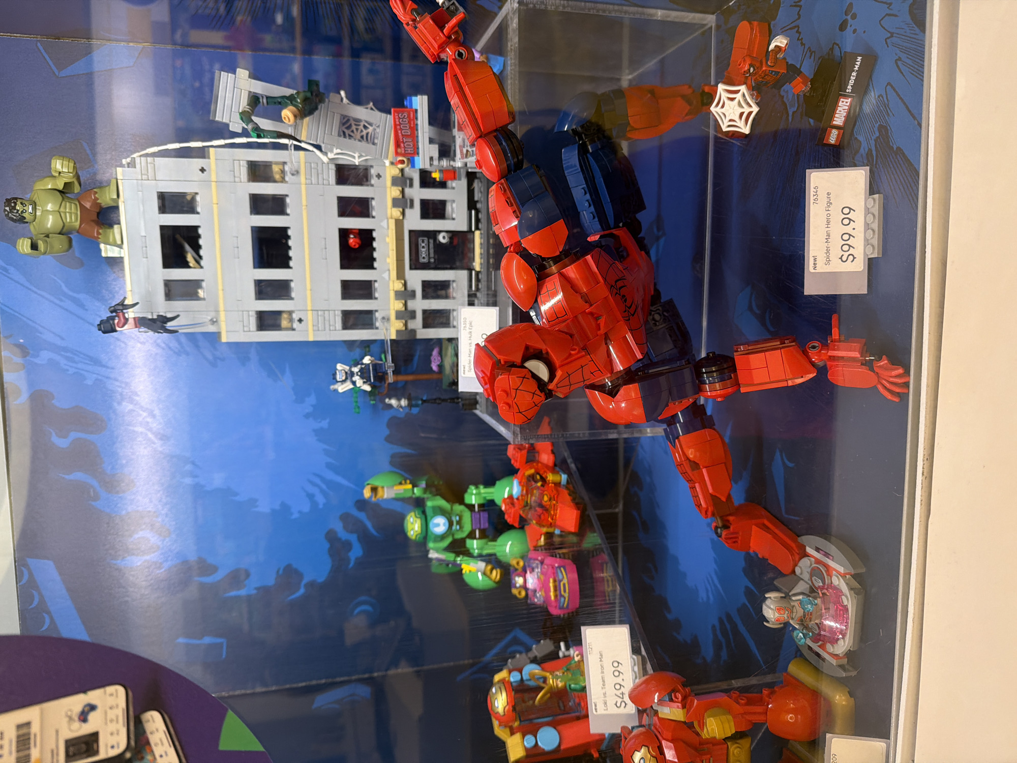 The image displays a retail store window in Mission Viejo, United States of America, showcasing various LEGO Marvel superhero merchandise. The primary focus is a large LEGO Spider-Man figure in a dynamic pose, positioned prominently in the foreground. Behind Spider-Man, a LEGO building is depicted with several figures including the Hulk, Spider-Man, and other characters engaged in what appears to be a battle scene. To the left, smaller LEGO sets are visible, including a green mech suit, a pink vehicle, and a red vehicle with a Loki figure. A price tag for "Loki vs. Team Iron Man" is visible, indicating a price of $49.99. Further to the right, a price tag for "Spider-Man Hero Figure" is displayed with a price of $99.99, alongside a LEGO Marvel Spider-Man display. The background features a blue, comic-book style graphic with flame-like designs. The lighting suggests an indoor retail environment.
