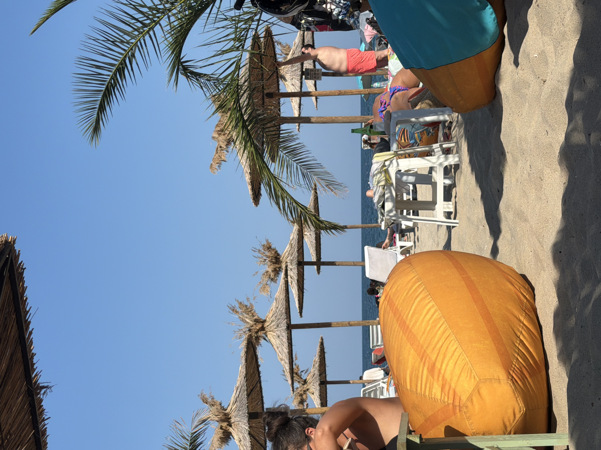 The image depicts a sunny day at a beach, likely in Varna, Bulgaria, given the context. In the foreground, a large, orange beanbag chair rests on the sandy beach, partially obscured by shadows. To the right, a turquoise object, possibly another beanbag or cushion, is also visible.
Behind the beanbag, several thatched umbrellas are lined up, providing shade. In the mid-ground, people are scattered across the beach, some lounging on chairs, others standing. One man in red shorts is seen standing near an umbrella, possibly adjusting it. A green bottle sits on a table between two lounge chairs. The sea stretches out in the background under a clear blue sky. Palm fronds from a tree overhang the scene from the right side. The overall atmosphere is one of a relaxed beach day with clear, warm weather.