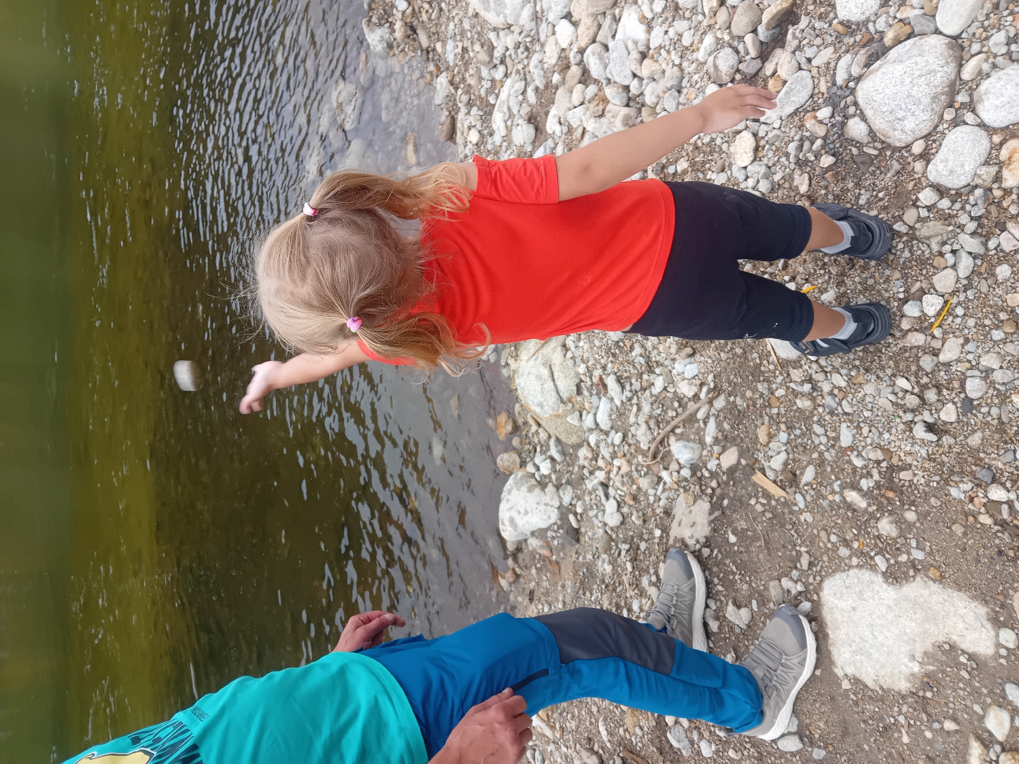 In Habovka, Slovakia, on a rocky riverbank, a young girl with blonde hair in pigtails is captured mid-action, throwing a stone into a calm, green body of water. She is wearing a bright orange off-the-shoulder shirt and black shorts. To her left, an adult, partially visible from the waist down, stands wearing a teal t-shirt and blue and gray athletic pants, along with grey athletic shoes. The ground is covered with a variety of smooth, grey and white stones, with a few larger rocks interspersed. The water's surface shows subtle ripples, indicating a gentle current.