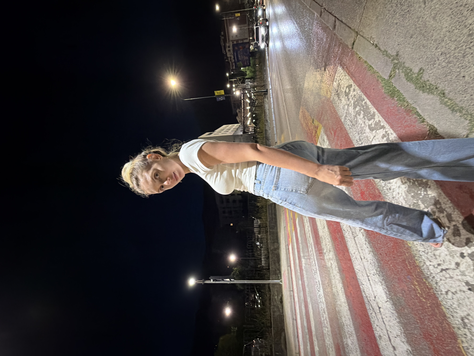A young woman with blonde hair in a bun stands on a wet pedestrian crossing at night. She is wearing a white ribbed t-shirt and loose-fitting blue jeans. The pedestrian crossing has alternating red and white stripes, with some yellow markings visible. Streetlights illuminate the scene, casting reflections on the wet asphalt. In the background, there are buildings, parked cars, and streetlights. The location is identified as Budva, Montenegro.