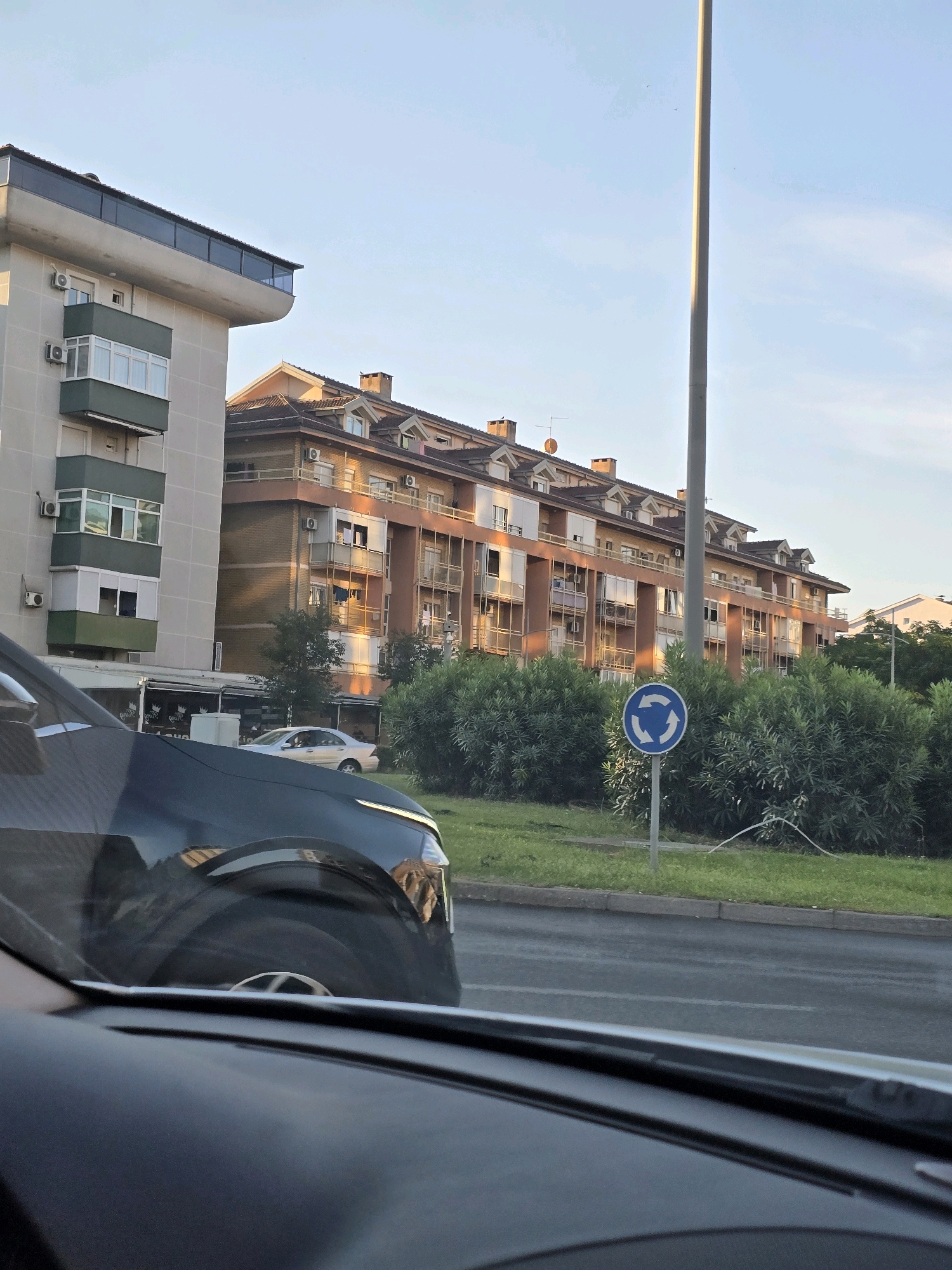 The image shows a street scene in Podgorica, Montenegro, as viewed from inside a vehicle. In the foreground, the dashboard and windshield of the car are visible. The exterior of a dark-colored car is partially seen on the left. Beyond the car, there are buildings with balconies and windows, and lush green bushes line the street. A traffic sign indicating a roundabout is prominently displayed on the right. The sky is clear and blue, suggesting it is daytime. The overall impression is of a typical urban environment in a Mediterranean setting.