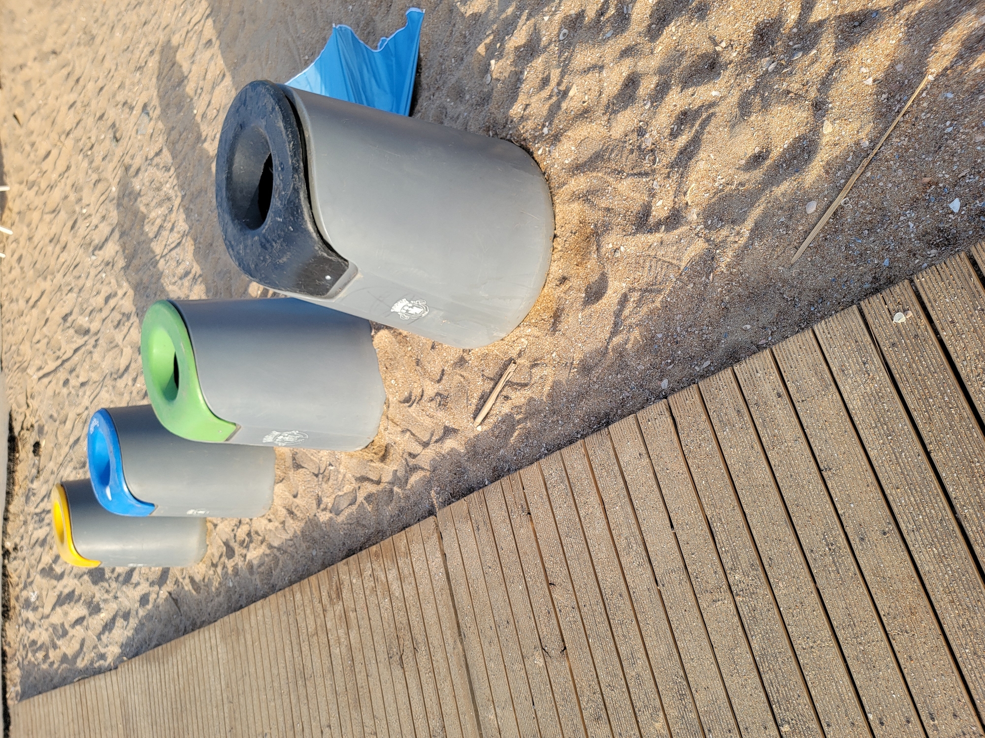 The image displays a close-up, overhead view of four colorful recycling bins placed on a sandy beach next to a wooden boardwalk. The bins are arranged in a staggered line, with the largest one at the top left and progressively smaller ones below it. The top bin is gray with a black opening. Below it, a gray bin has a bright green opening. The third bin, also gray, features a blue opening, and the smallest, bottom bin is gray with a yellow opening. A piece of blue plastic, possibly a beach umbrella or mat, is visible behind the top bin. The sand is coarse and speckled with small shells and pebbles, and dappled sunlight casts shadows across the scene. The wooden boardwalk runs diagonally from the bottom right of the frame, its planks worn and weathered. The scene is set in Quarteira, Portugal, and the lighting suggests it is a sunny day, likely in the late afternoon given the long shadows. There is no visible text in the image.