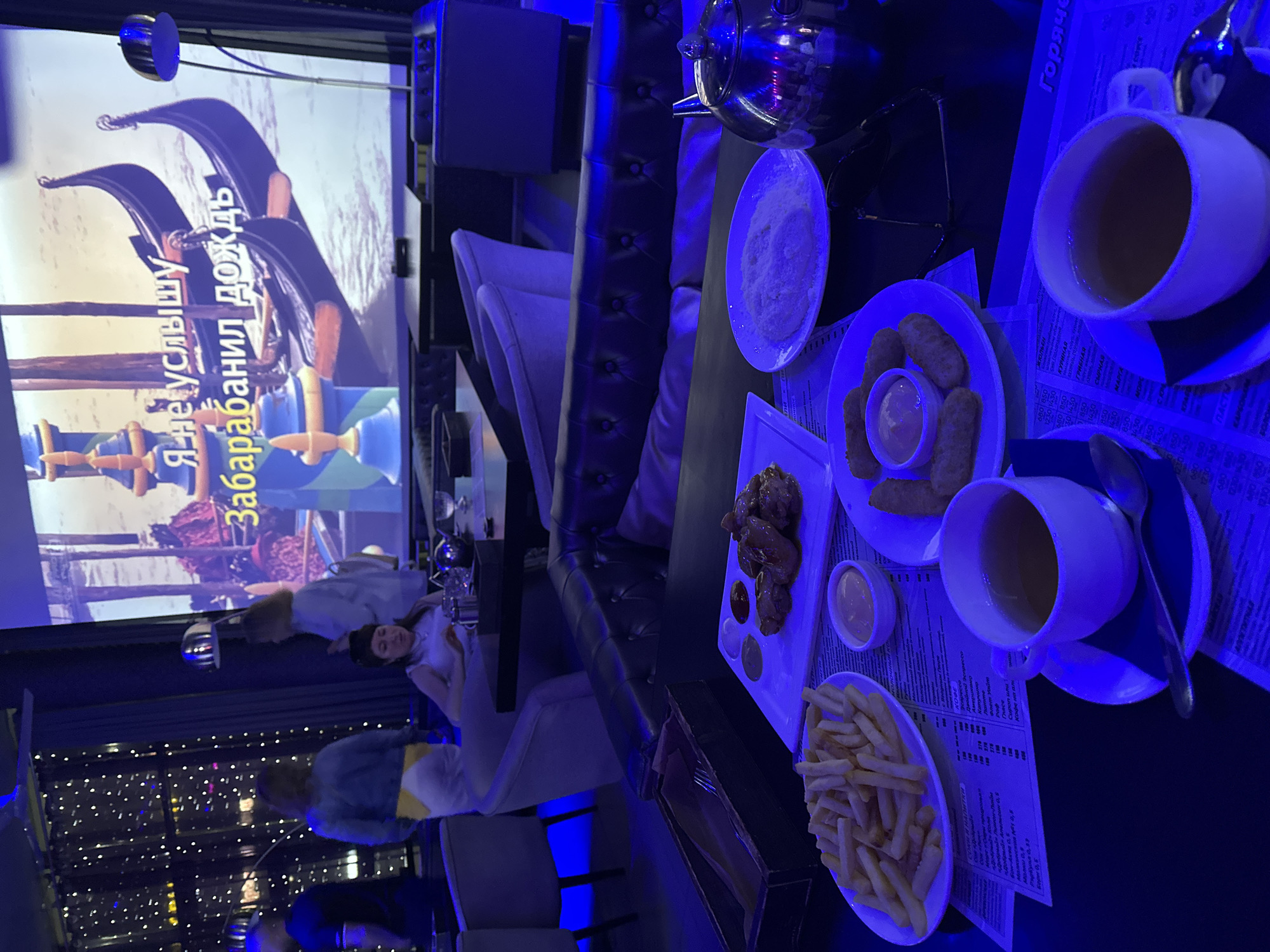 The image depicts an indoor scene, likely a restaurant or bar, bathed in blue ambient lighting. In the foreground, a dark table is set with two cups of tea, a plate of french fries, a plate of chicken wings with dipping sauces, a plate of what appears to be grated cheese or breadcrumbs, and a teapot. A menu is visible on the table. In the background, a projection screen displays an image of gondolas with Russian text overlaid. Several people are present in the background, some standing and some seated, with a curtain or decorative lighting element behind them. The overall atmosphere suggests a casual dining or entertainment setting. The location is stated to be the city of Petropavlovsk-Kamchatsky, Russia.