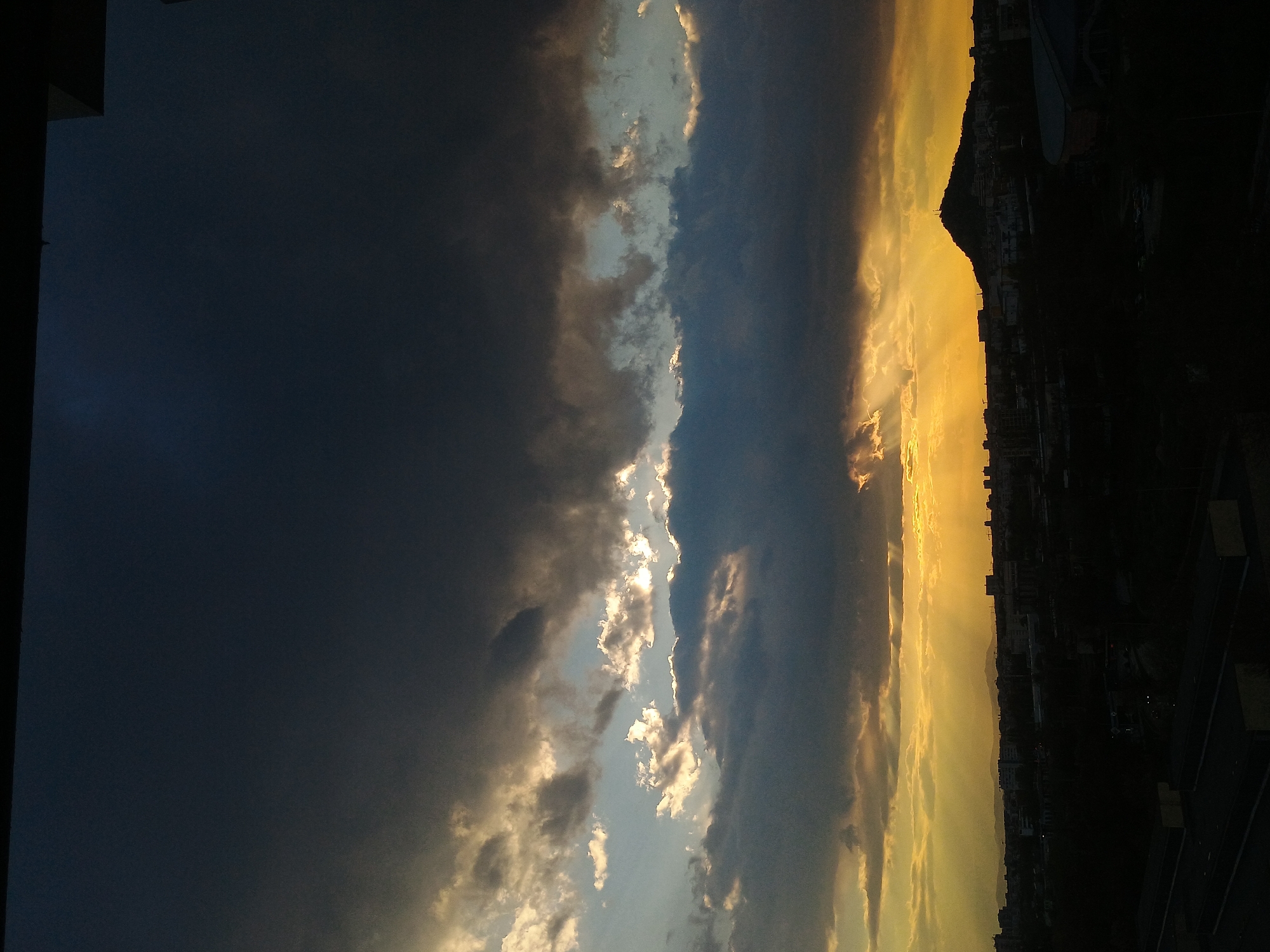The image shows a sunset view in Plovdiv, Bulgaria. The sky is divided into three distinct parts: a dark, almost black area on the left, a central area with dramatic clouds illuminated by the setting sun, and a bright, golden area on the right where the sun is setting.
The right side of the image reveals a city skyline silhouetted against the bright sky. Details of buildings and structures are visible, with a mountain or hill rising prominently in the background. Below the skyline, there is what appears to be a cityscape with buildings and possibly roads.
The overall atmosphere is dramatic and suggestive of the transition from day to night, with the contrast between the dark, cloudy sky and the bright sunset creating a stark visual impact. The photograph seems to be taken from an elevated position, possibly from a high-rise building, allowing a wide view of the city and the surrounding landscape.