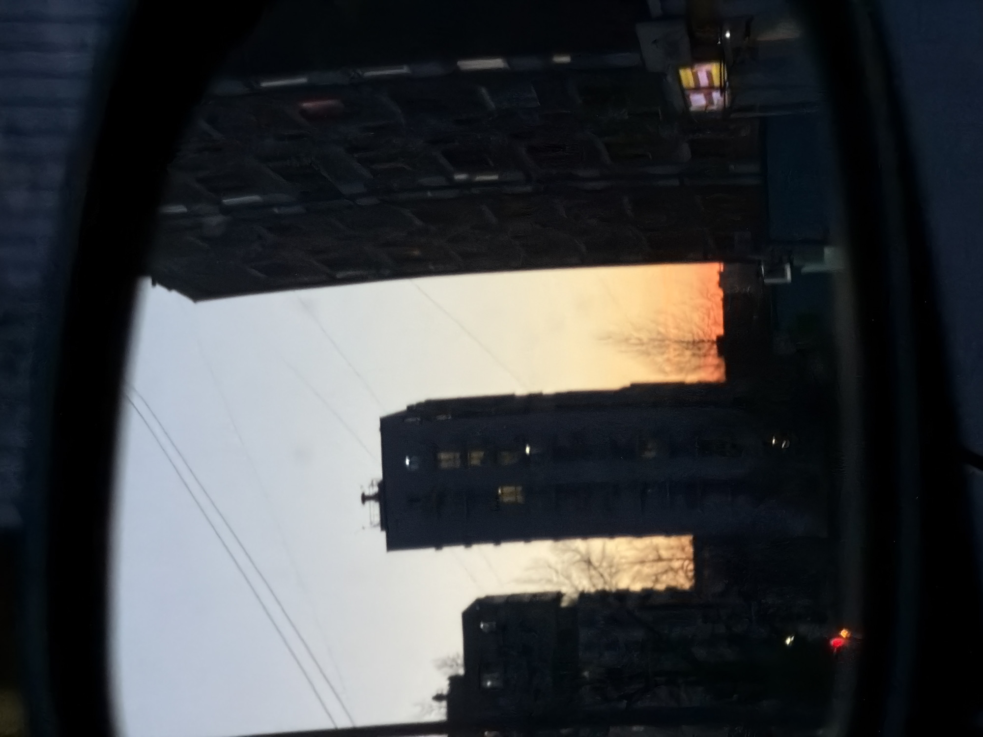 The image, likely a photograph taken in Izhevsk, Russia, appears to be a reflection in a rearview mirror. The reflection shows a city scene with several multistory buildings. The sky is illuminated by the colors of a sunset. The buildings are mostly dark silhouettes, though some windows show light. Power lines are visible across the sky. The mirror's frame is evident around the reflection, and the background outside of the mirror is dark.