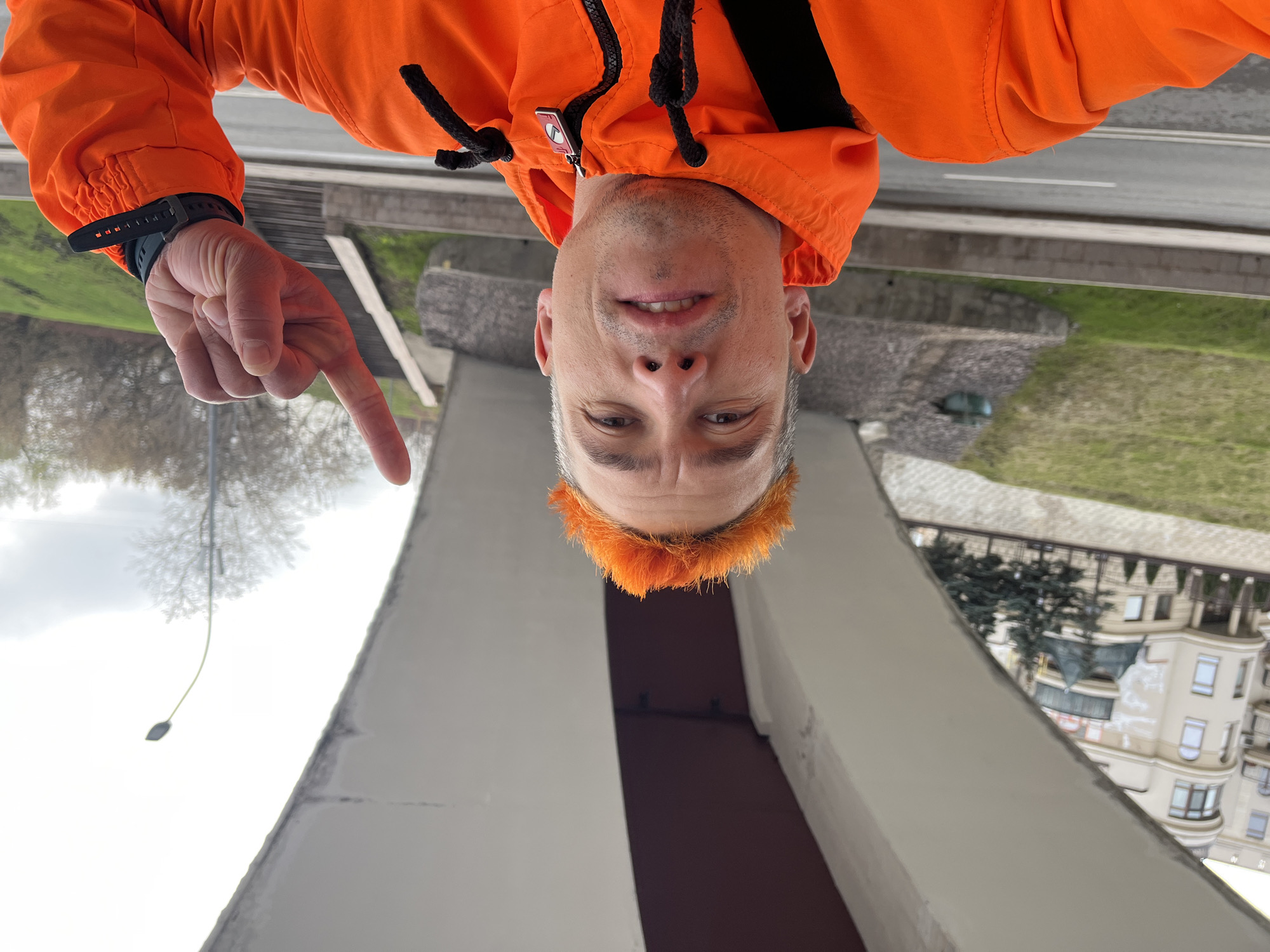 A person in an orange jacket, facing downward in the frame, is pointing towards a tall, white and brown structure. The structure appears to be a modern architectural element, possibly part of a bridge or monument. The background features a building with multiple windows and architectural details, suggesting an urban setting. Other objects, such as a streetlight and trees, are visible in the scene. The city is Moscow, Russia.