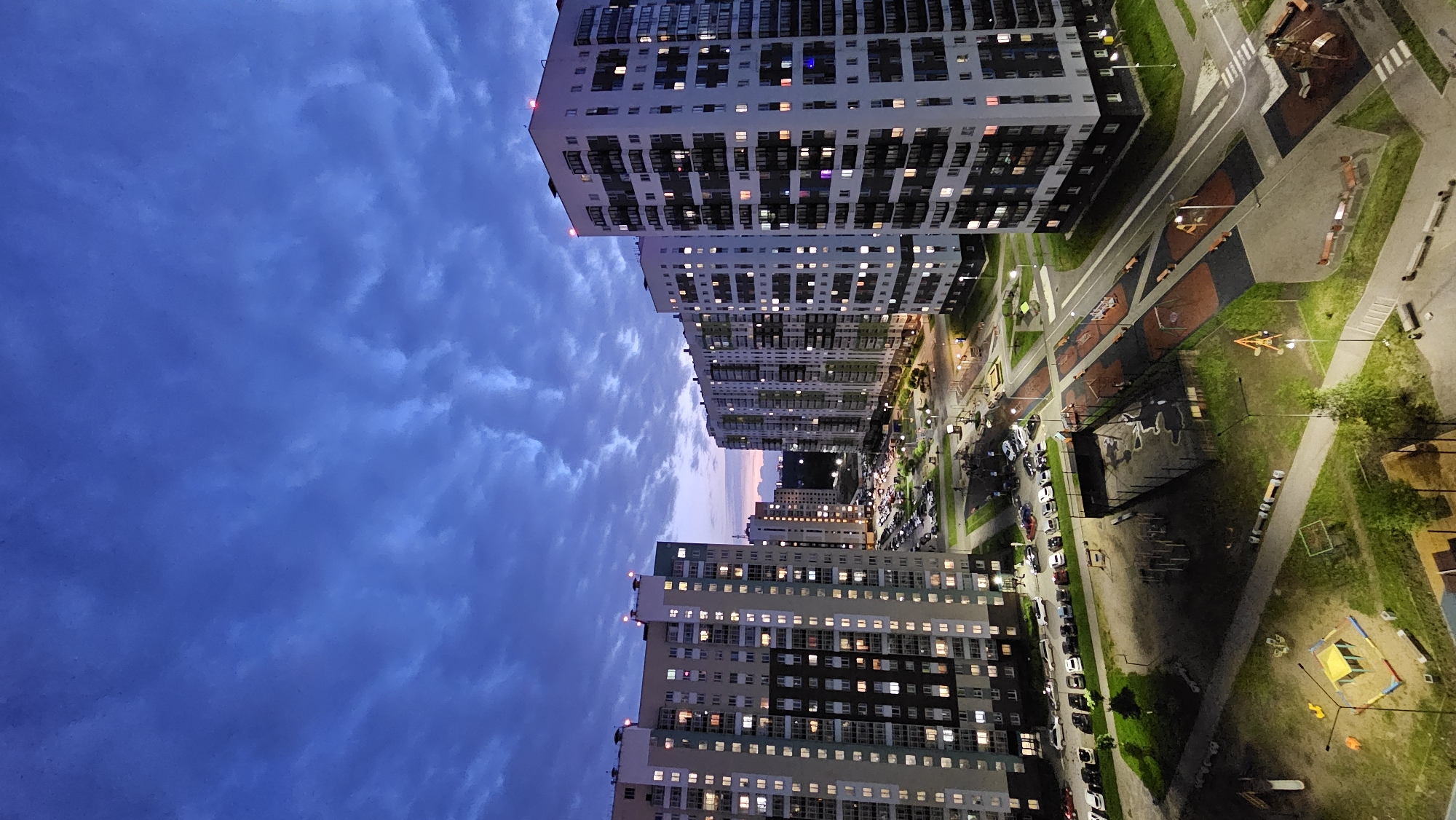 The image depicts a twilight view of a residential complex in Yekaterinburg, Russia. Several tall, multi-story apartment buildings dominate the scene, their windows illuminated with warm light. The architecture suggests a modern urban development. A parking lot filled with cars is visible between two of the buildings, indicating a populated area. Below the buildings, there are courtyards with children's playgrounds, including swings, slides, and sandboxes, some of which are dimly lit by streetlights. The sky above is a deep blue, characteristic of dusk, with scattered clouds that are beginning to catch the last rays of sunlight on the horizon. The overall impression is that of a quiet evening in a contemporary urban environment.
