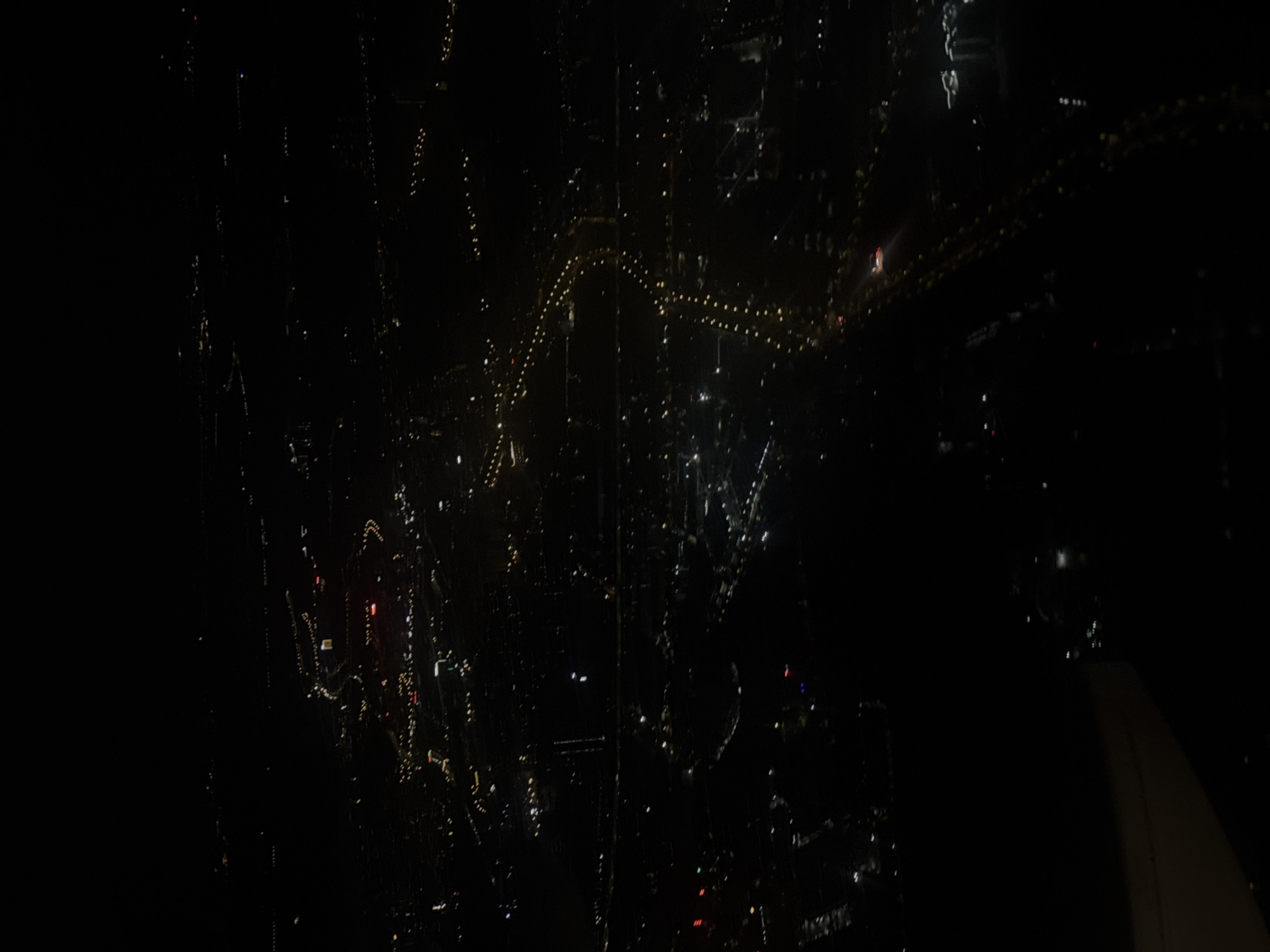 The image presents a dark, high-angle view of a city at night, likely seen from an airplane or a tall structure. The dominant feature is a sprawling pattern of numerous small, illuminated lights against a black backdrop, characteristic of an urban landscape after dark.
A prominent element in the lower right portion of the frame is the beige-colored wing of an aircraft, confirming an aerial perspective. The scene is viewed through a window, which exhibits noticeable streaks, smudges, and possibly water droplets or condensation, creating a textured overlay that distorts and diffuses some of the distant lights.
Among the scattered city lights, a distinct, winding arc of golden-yellow lights is visible, suggesting an illuminated bridge, a major roadway, or a decorative installation. Other lights appear as tiny, shimmering points or short, blurry streaks, their forms influenced by the window's imperfections. The time of day is clearly nighttime. Given the context of Huangpu District, China, these are likely the vibrant night lights of Shanghai. There is no discernible text within the image.