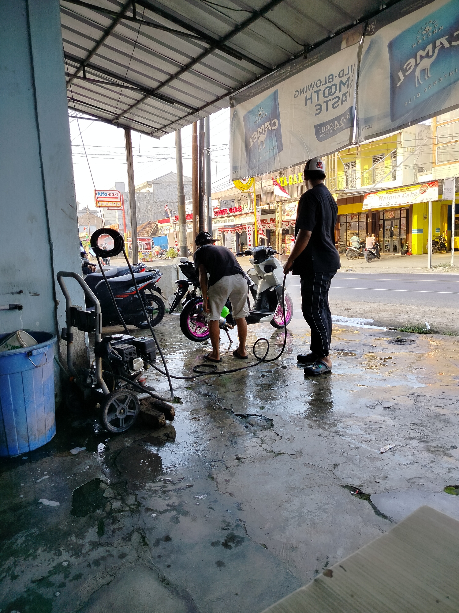 Two men are washing a white motorcycle at an outdoor car wash. The car wash is covered by a corrugated metal roof supported by metal poles. The floor is wet concrete with puddles of water. To the left is a blue plastic bin and a pressure washer. The men are wearing casual clothing and flip-flops. The motorcycle has a pink rim on the back wheel. In the background, there are several shops, including a brightly colored yellow building with a sign that says "Halona Cake." There is also a red and white Indonesian flag visible. The sky is overcast. The scene is likely located in Braja Asri, Indonesia.