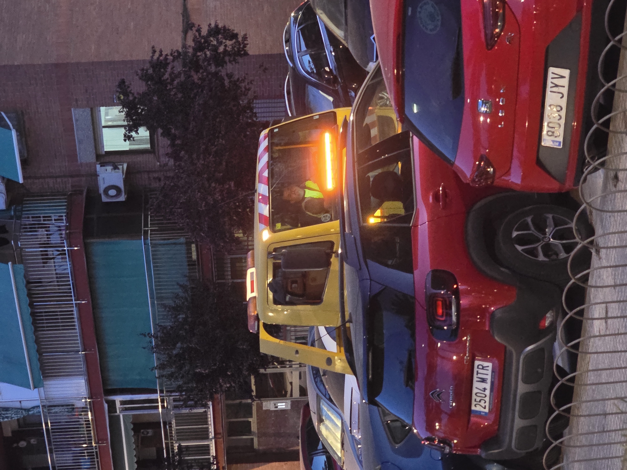The image depicts a street scene in what appears to be Cornellà de Llobregat, Spain, given the license plate formats. A yellow tow truck is parked on the street, its emergency lights activated. A person wearing a high-visibility vest can be seen inside the tow truck. A red Citroen C3 Aircross, license plate 2504 WTR, is parked in front of the tow truck, with another red car, license plate 8038 JYY, parked adjacent to it. A building with several stories is visible in the background, with blinds covering some of the windows. An air conditioning unit is mounted on one of the walls. A tree stands near the building. A fence is visible in the foreground.