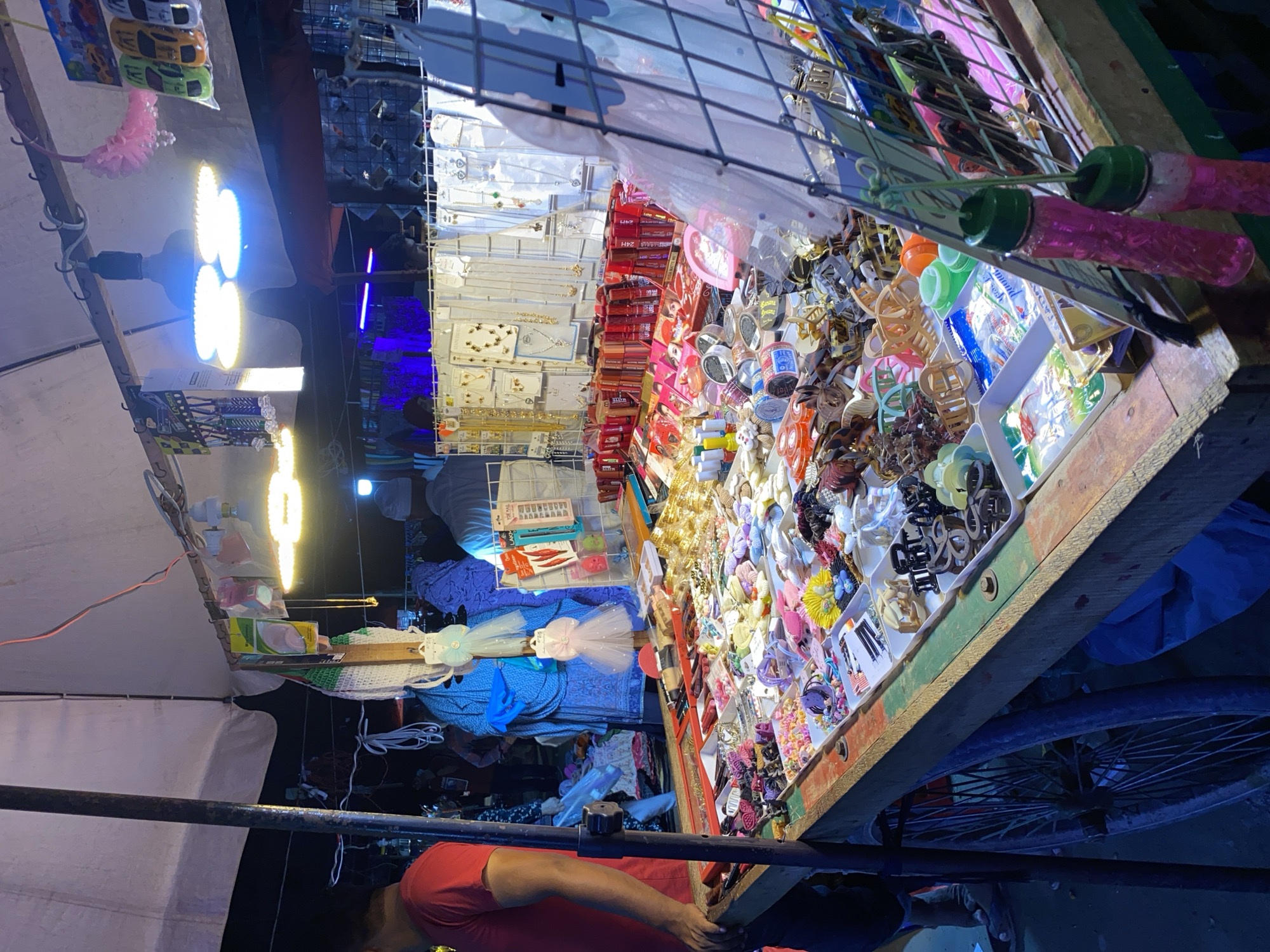 This image captures a brightly lit street vendor's stall at night, characteristic of a bustling market in Dhaka, Bangladesh. The stall, likely a mobile cart built on a bicycle frame (a wheel is visible underneath), is overflowing with a diverse array of merchandise.
The display is illuminated by several powerful LED lights, including large circular bulbs emitting both cool blue and warm yellow light, as well as linear light strips, creating a vibrant glow against the dark background.
The main subject is the merchandise itself, which includes a vast collection of hair accessories such as colorful plastic and metal hair clips, claw clips, and hair ties, meticulously arranged in white trays. Adjacent to these are various jewelry items like necklaces, earrings, and bracelets, presented on hanging cards. Further along the stall, small packaged cosmetics or beauty products, some in red boxes marked "24H," are visible. Hanging elements include a packet of toy cars and distinctive pink bubble wands with green caps.
In the lower right quadrant, a person wearing a red shirt is partially visible, leaning near the stall, possibly tending to it or observing the goods. The overall scene suggests a lively commercial activity taking place after dusk, with a dense concentration of affordable goods for sale.