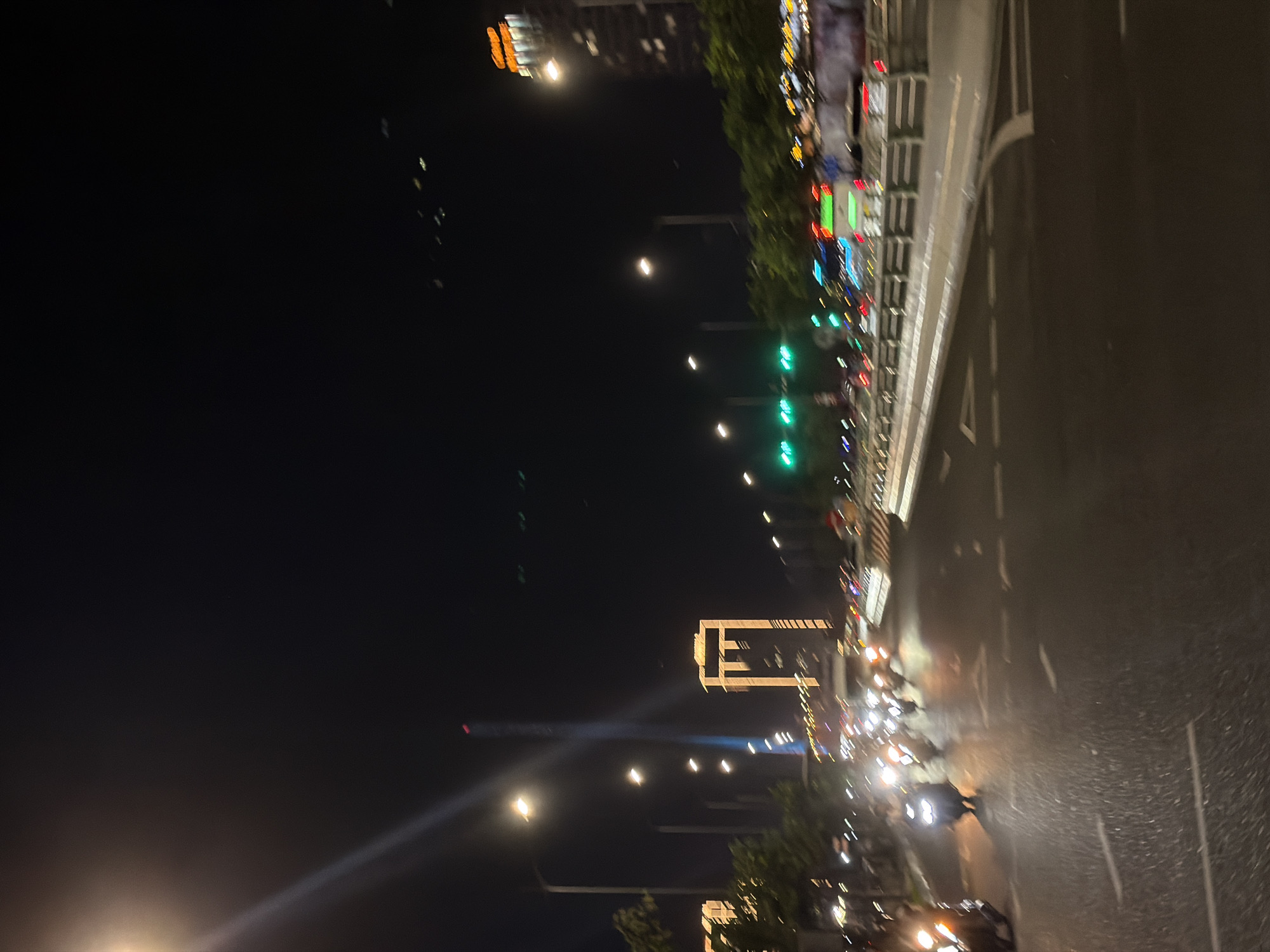 The image depicts a nighttime scene on a multi-lane road in Đà Nẵng, Vietnam. The road is wet, reflecting the lights from street lamps, traffic signals, and vehicles. A prominent feature in the background is a tall, illuminated skyscraper that appears to be the Landmark 81, with a distinctive illuminated outline. The foreground shows a group of motorcycles and scooters traveling on the road, their headlights creating streaks of light due to motion blur. Some vehicles in the distance also show similar motion blur. The environment appears urban, with trees and other buildings visible along the sides of the road. The sky is dark, indicating it is night. The overall impression is a busy urban thoroughfare at night, characterized by vehicle lights and illuminated architecture.