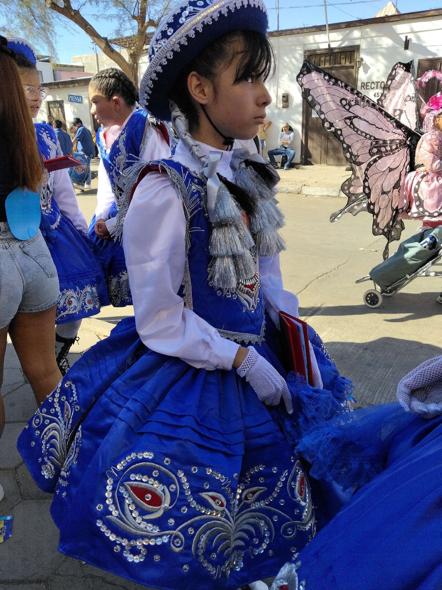 In Pozo Almonte, Chile, a young girl in a blue and silver traditional costume, complete with a wide-brimmed hat, stands in the foreground. Her costume features intricate silver embroidery and red accents on the bodice and skirt. She wears white lace gloves and holds a red book. Behind her, other children in similar costumes are visible. To the right, a woman in a pink costume with large butterfly wings is also present. The scene appears to be outdoors, on a street, with buildings and trees in the background. The overall impression is of a cultural event or parade.