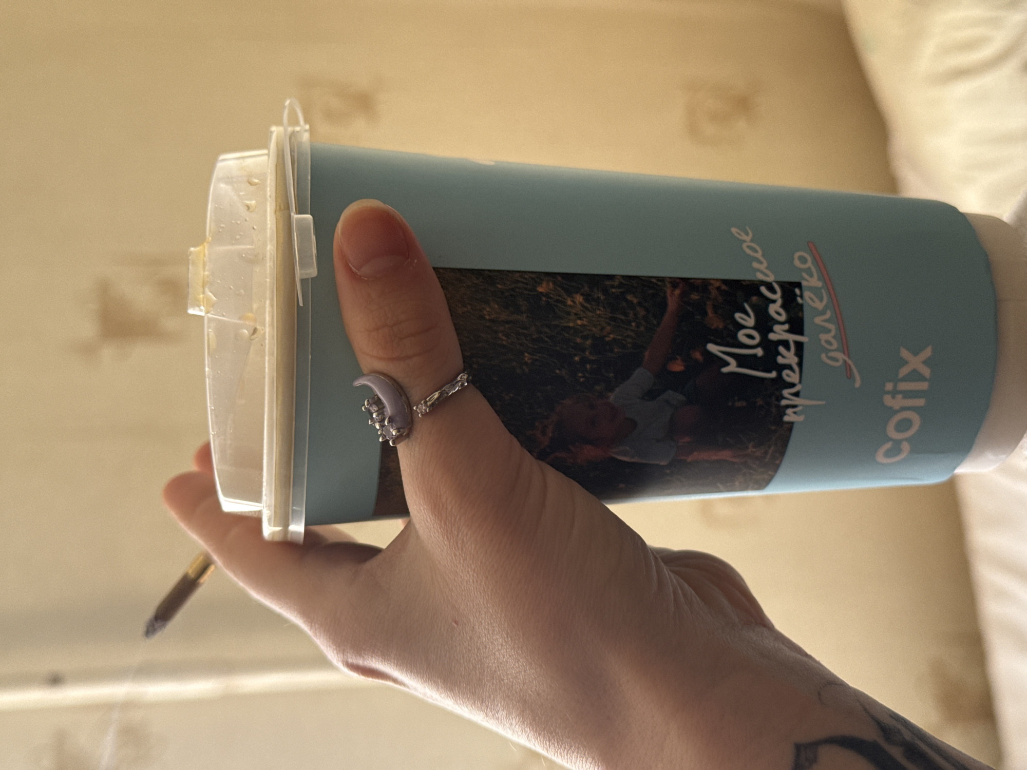 A hand holds a light blue Cofix brand coffee cup. The cup's label contains an image of children in nature. The text "Мое прекрасное далеко" is printed on the cup. A lit cigarette or joint is positioned in the hand holding the cup. The person is wearing a silver ring on their thumb. The background appears to be a room, possibly in an apartment or house. The location is Moscow, Russia.
