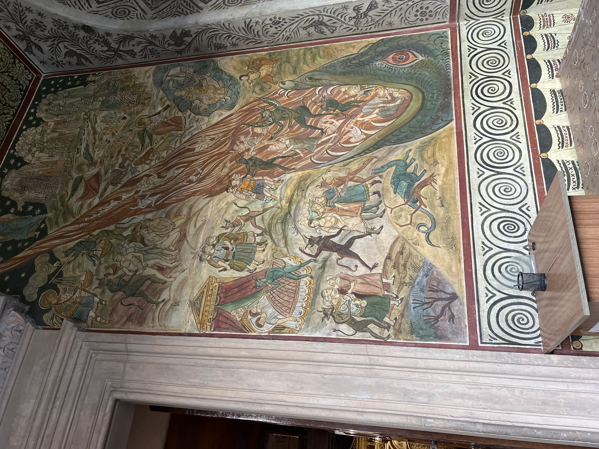The image shows a fresco on the wall of a building, likely a church, in Constanța, Romania. The fresco depicts a chaotic scene with numerous figures engaged in various activities. In the upper portion, a large, fiery plume descends from the sky, engulfing some of the figures. Angels and demonic figures are present, with one angelic figure appearing to weigh souls. Below this, there are scenes of people in beds, some being disturbed by demonic beings, and groups of figures interacting with each other and with demons. A large, serpentine creature with prominent eyes dominates the lower right portion of the fresco, seemingly consuming or threatening the figures. The lower part of the wall features a decorative border with spiral patterns and stylized motifs. The fresco style is traditional and appears to be old.