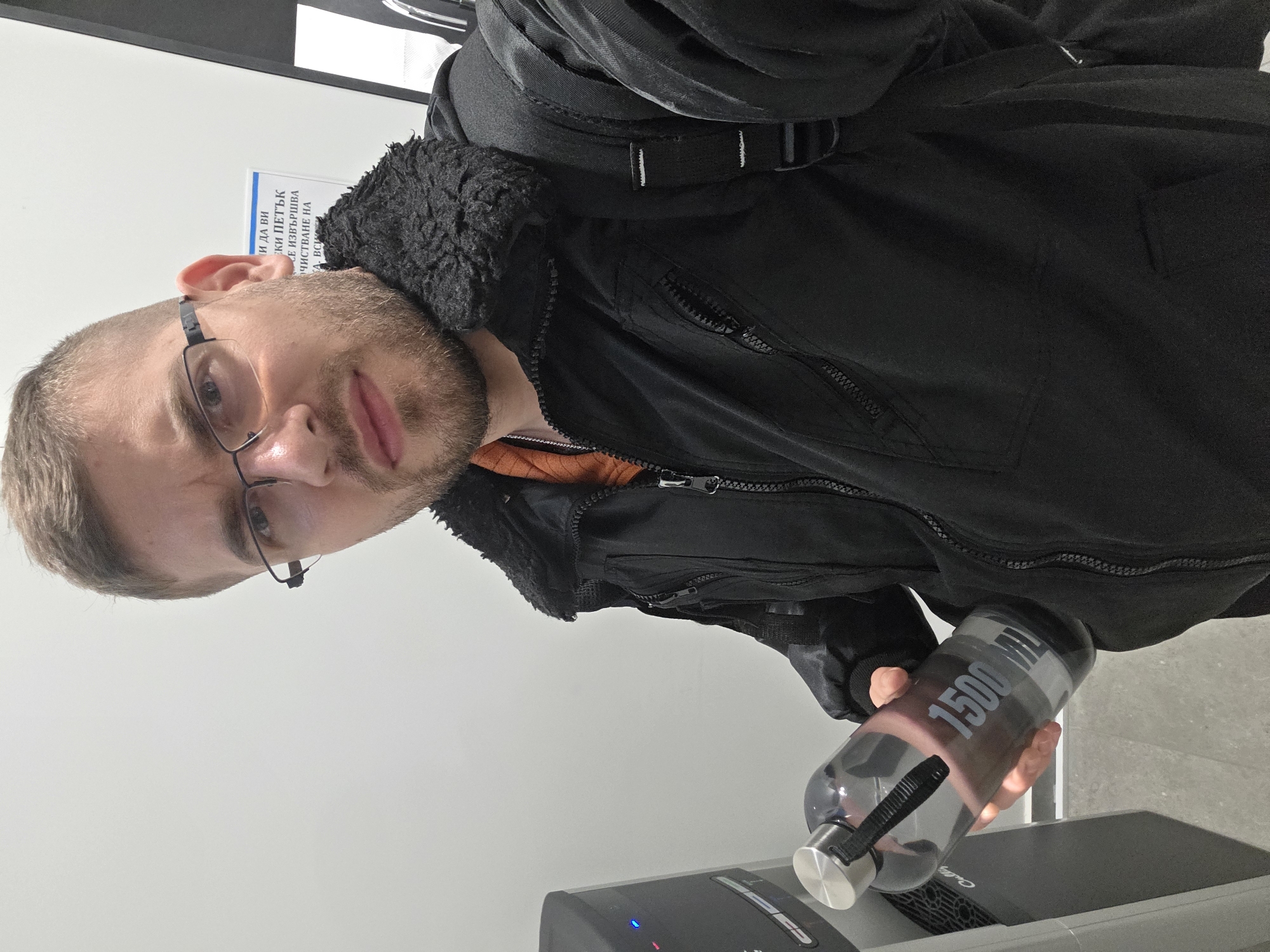 A man with glasses, a beard, and wearing a black jacket with a fur collar and a backpack stands indoors in Plovdiv, Bulgaria. He is filling a 1500ml water bottle at a water dispenser. A sign with Bulgarian text is partially visible on the wall behind him.