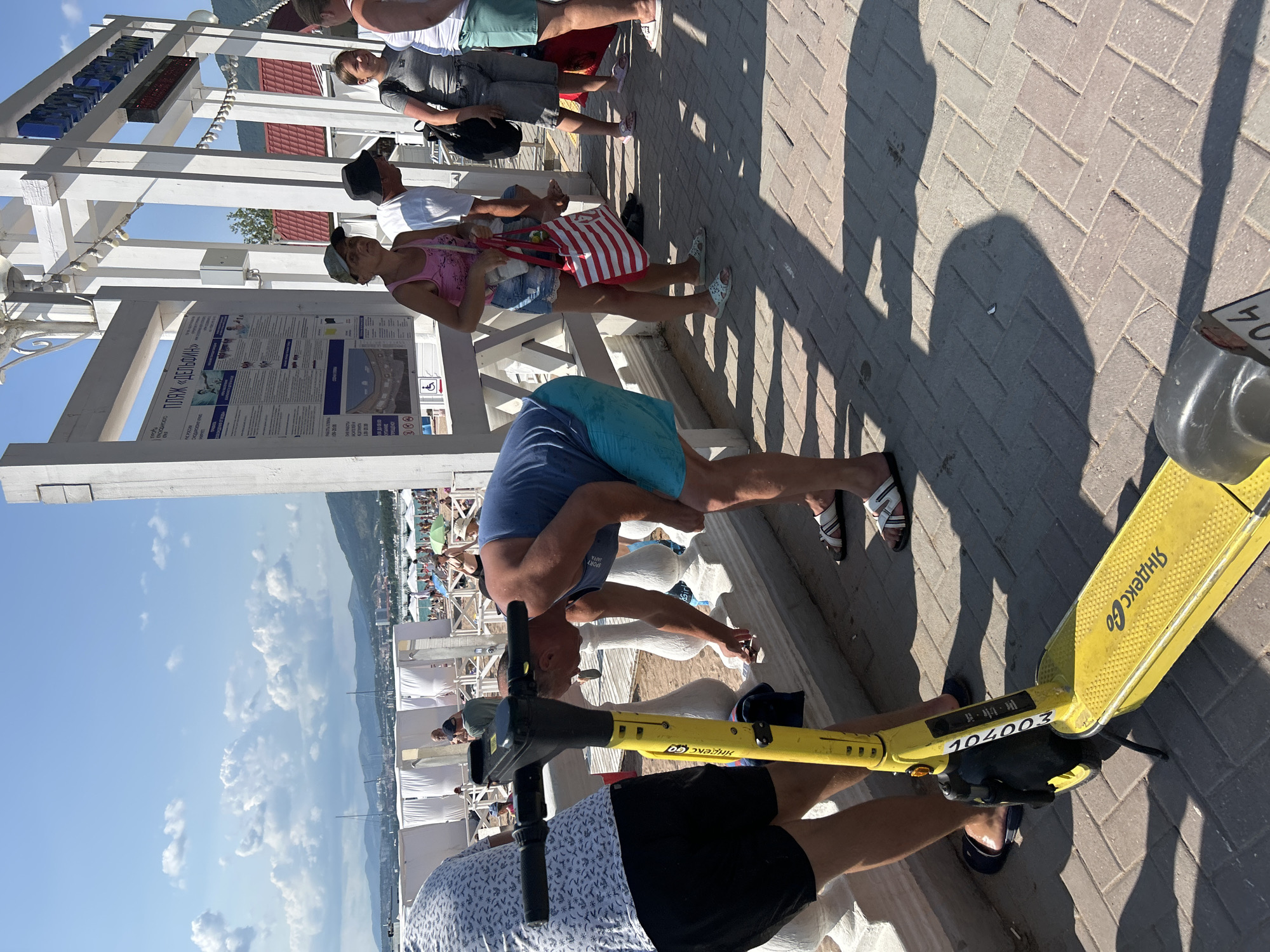 The image depicts a bright, sunny day at a beach boardwalk in Gelendzhik, Russia. The foreground features a yellow electric scooter with the inscription "Яндекс Go". To the left, a person wearing a patterned t-shirt and black shorts stands with their back to the camera, facing the scooter. In the center, a man in a blue tank top and blue shorts is bending down, seemingly interacting with the ground near a white railing. Behind him, several other people are visible. To the right, a woman in a pink tank top and denim shorts carries a red and white striped bag. Next to her, a man in a dark hat and a white shirt, and a young woman in a grey t-shirt, are also present. In the background, white beach cabanas, beachgoers, a sea, and a mountainous landscape under a blue sky with clouds can be seen. A large white structure with information boards is also prominent on the right side of the image.