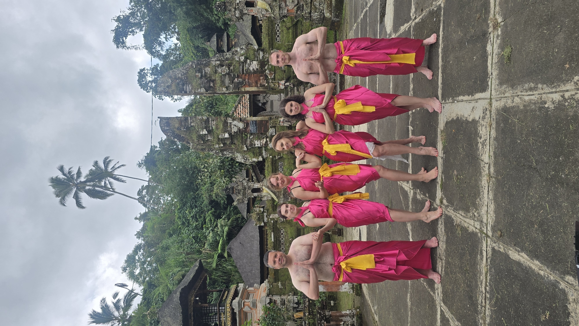 A group of six people is lined up posing in front of a temple structure in Tegallalang, Indonesia. Two men stand at either end of the line, shirtless, wearing hot pink and yellow sarongs. Between them, four women stand wearing pink dresses tied with yellow belts or sashes. Everyone has their hands pressed together in a prayer-like gesture, facing the camera. The group stands on a cracked grey concrete surface. Behind them is a jungle and a multi-level temple structure made of grey stone and covered in moss and plants. The sky is overcast and the image appears to be rotated 90 degrees counterclockwise.