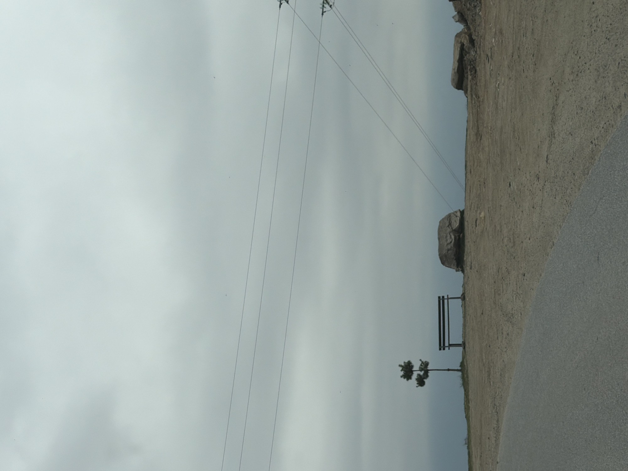 A landscape featuring an overcast sky, power lines, a rocky hillside, and a road. A bench and a small tree stand at the edge of the hill, near the road. The image might be taken in a rural or semi-rural area, potentially near Leshten, Bulgaria.