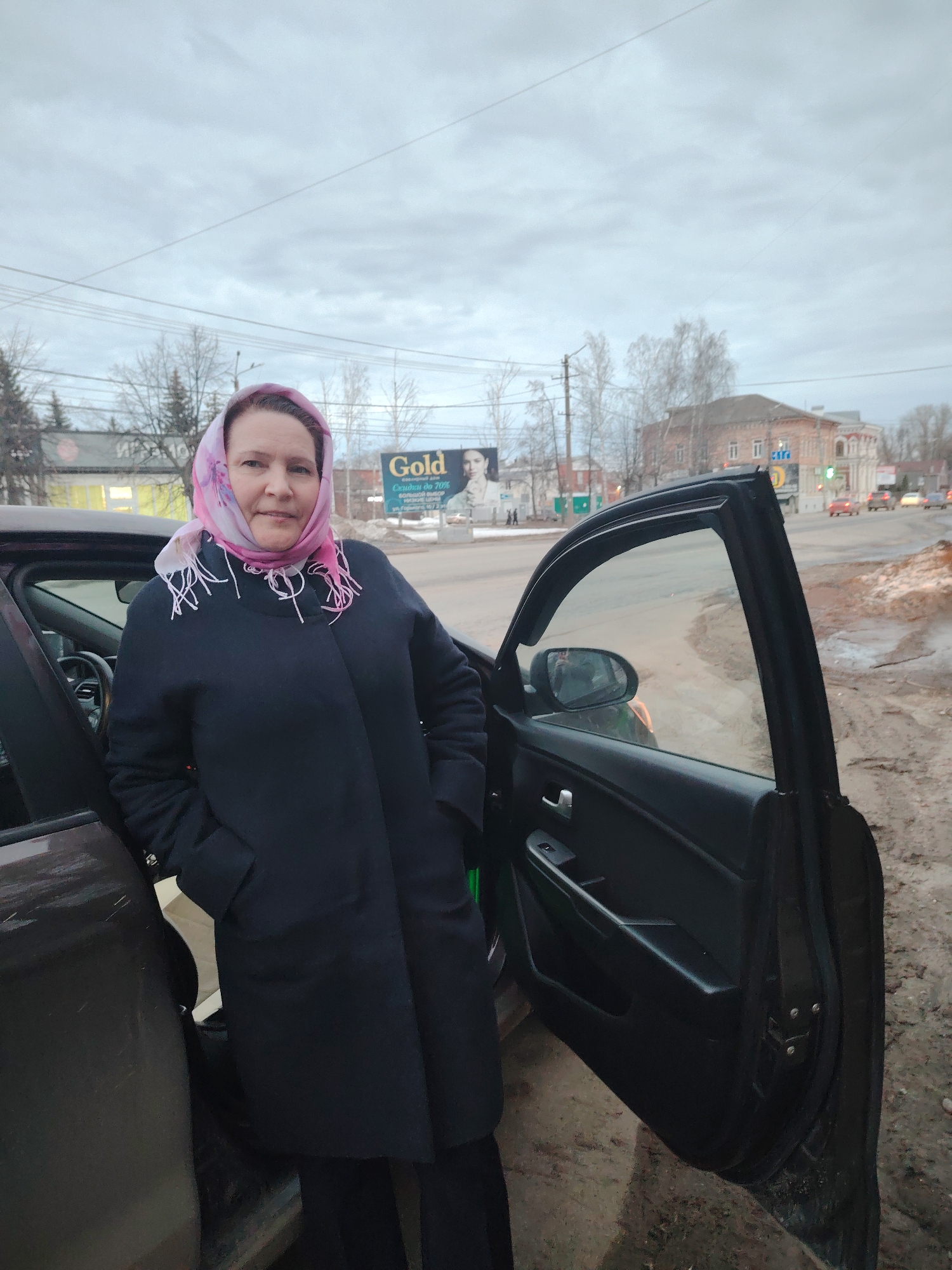 A middle-aged woman stands beside a car on a cloudy day in Sarapul, Russia. She wears a dark coat and a pink headscarf. Her hands are in her pockets. The car door is open. In the background are buildings, trees without leaves, and a "Gold" advertising billboard. Cars are visible on the street. The ground beside the car appears muddy.