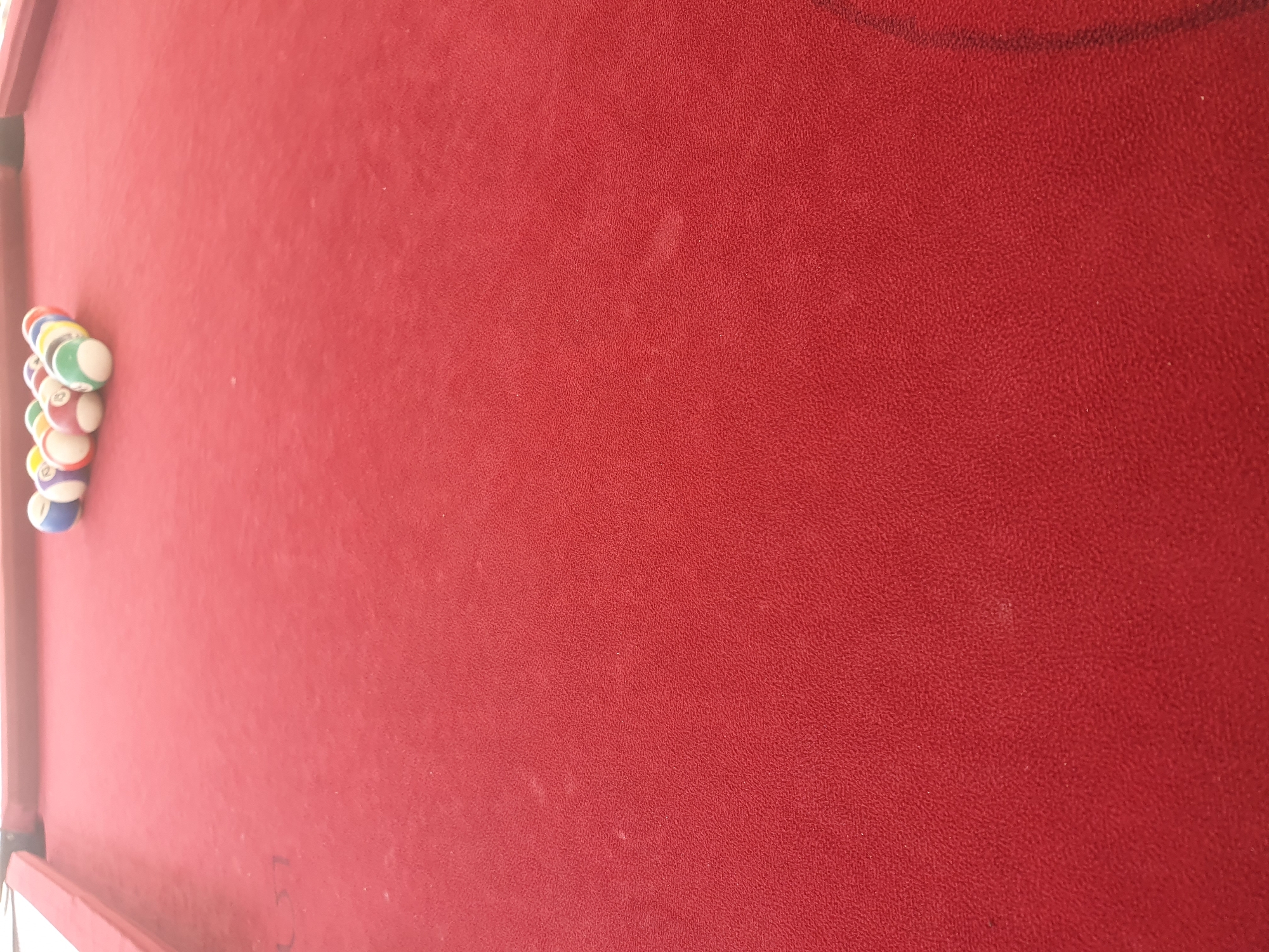 A close-up, low-angle shot shows a rack of pool balls on a red felt billiard table. The balls are arranged in a triangular formation, with the apex ball at the front. The image focuses on the texture of the felt, which appears slightly worn and dusty, with some lighter specks visible. The pool balls are a mix of solid colors and stripes, with numbers ranging from 1 to 15. The background is a blur of red, suggesting the rest of the table extends out of frame. There are no people or any other objects visible in the image. The scene is indoors, likely in a casual setting such as a bar or game room. The location is not specified in Lokoja, Nigeria.