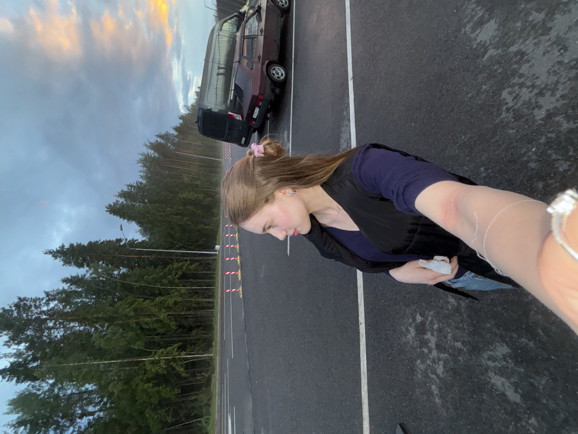The image shows a young woman standing on an asphalt surface, likely a road or parking lot, near a line of trees. She is wearing a dark vest over a blue top and jeans. Her right arm is extended, holding a white object, possibly a tissue or cloth. Her hair is pulled back into a bun with a pink accessory. In the background, there are two vehicles: a dark van and a maroon sedan. The sky is cloudy with hints of sunset colors. The scene appears to be outdoors in a somewhat rural or suburban setting, potentially near Arfa, Russia, given the mention of the location. The overall lighting suggests it is late afternoon or early evening.