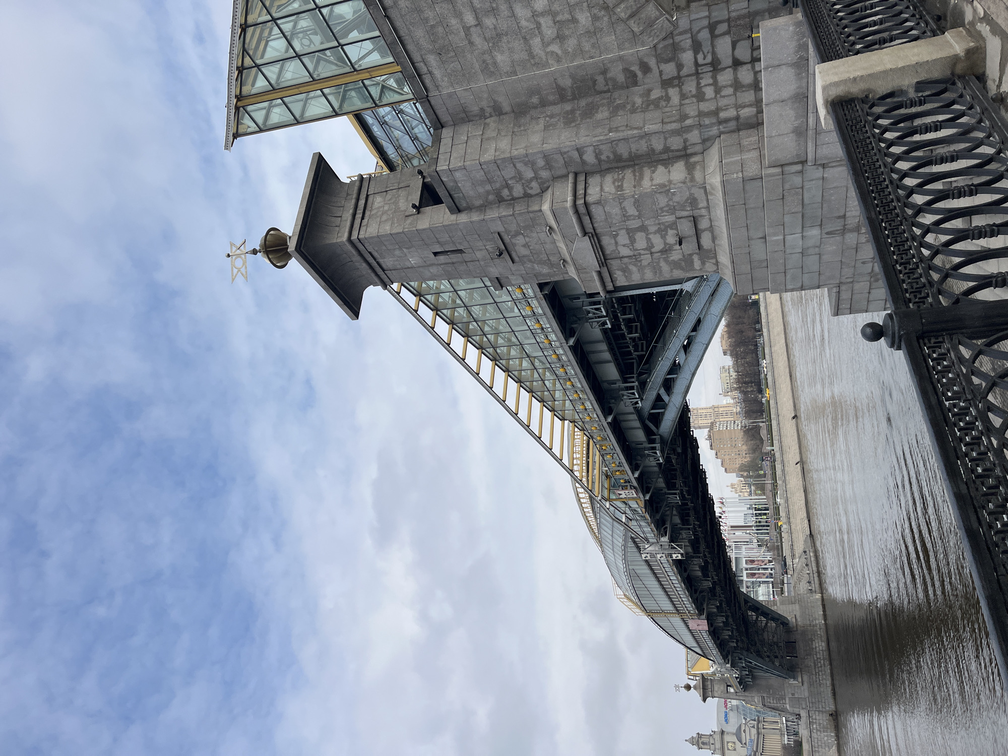 The image shows a view of the Borodinsky Bridge in Moscow, Russia. The bridge is made of grey concrete and steel, with a glass and steel pedestrian walkway running alongside the roadway. A decorative metal railing is visible on one side of the bridge, adjacent to the river. The Moscow River runs below the bridge, with buildings and structures visible on the riverbank. The sky is cloudy.