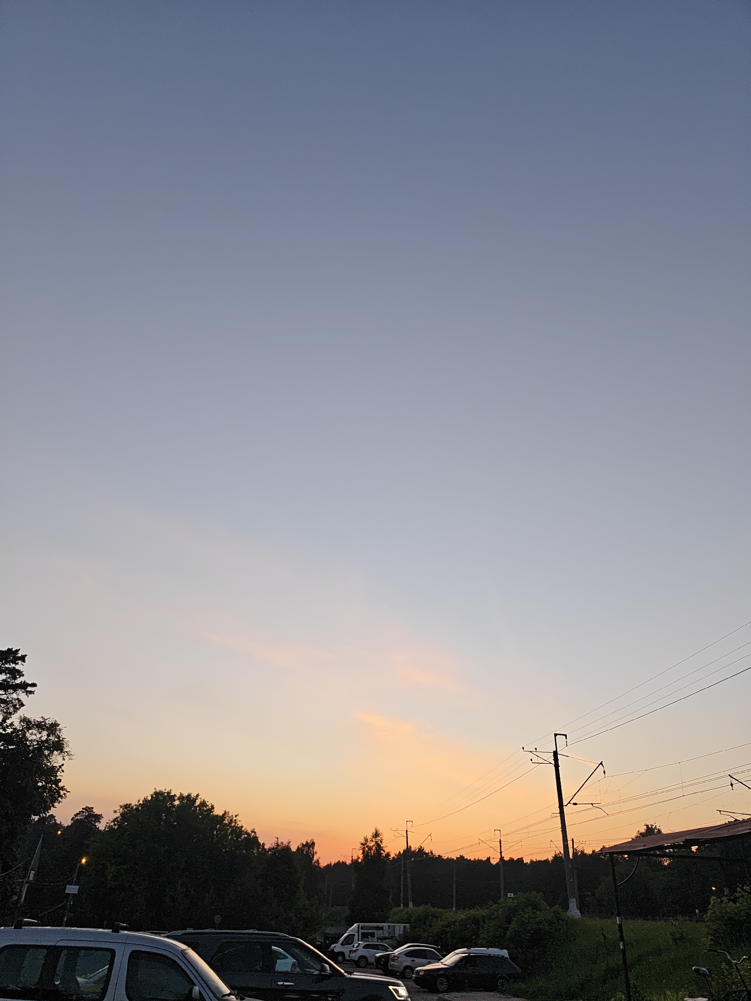 The image captures a sunset scene in Kratovo, Russia, characterized by a gradient sky and a cluttered suburban landscape. The upper portion of the sky transitions from a pale desaturated blue-grey to a soft orange and pink glow at the horizon, indicative of late evening light. The lower half of the image features a line of dense, dark green trees and shrubs silhouetted against the setting sun, forming a backdrop. In the immediate foreground, a silver-grey minivan or SUV is partially visible on the bottom left, parked alongside other dark-colored vehicles in what appears to be a parking area. Multiple tall utility poles with complex overhead electrical wires traverse the right side of the frame, extending into the distance. On the far right, a dark structure with a corrugated roof is partially visible. A faint artificial light source illuminates a small area within the trees on the left. No people are visible, and no discernible actions are taking place. The scene suggests a semi-urban environment near a forested area, possibly adjacent to a railway line due to the prominent electrical infrastructure.