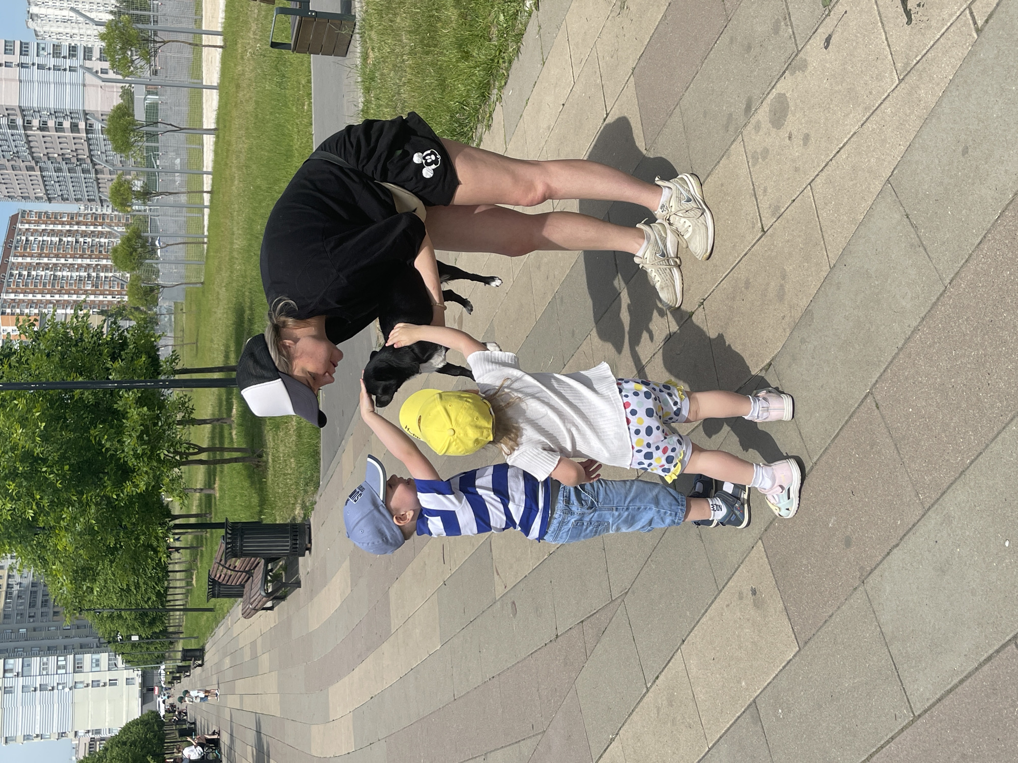 This image captures an outdoor scene on a paved walkway within a modern urban park, likely in the residential complex "LIFE-Botanichesky Sad" in Moscow, Russia, identifiable by the distinctive striped high-rise buildings in the background. The sky is clear and blue, indicating bright daylight conditions.
In the foreground, a woman, two young children, and a small black dog are interacting. The woman, positioned on the right and bending forward, wears a black short-sleeved top, black shorts with a white Mickey Mouse emblem on the left thigh, white athletic shoes, and a black and white baseball cap. She holds the small black dog in her arms, supporting its front paws with her left hand and its hindquarters with her right.
To the woman's left, a young boy, wearing a blue baseball cap, a white and blue horizontally striped t-shirt, and blue denim shorts, stands facing the dog. His right hand is extended, gently touching the dog's head. Next to him, a younger girl, dressed in a yellow baseball cap, a white short-sleeved top, and white shorts with yellow, blue, and red polka dots, also reaches out towards the dog with her right hand, near its body. Both children appear to be interacting with the dog under the woman's supervision.
The paved walkway consists of rectangular and curved light and dark grey interlocking tiles. To the right of the walkway is a patch of green grass with several small, newly planted trees protected by metal fences. In the middle ground, along the left side of the walkway, are several wooden park benches with black metal frames and associated black trash bins. Further down the walkway, more people are visible in the distance, some walking, others seated. Tall, multi-story residential buildings with varying architectural styles, including one with prominent horizontal blue and white stripes, dominate the background.