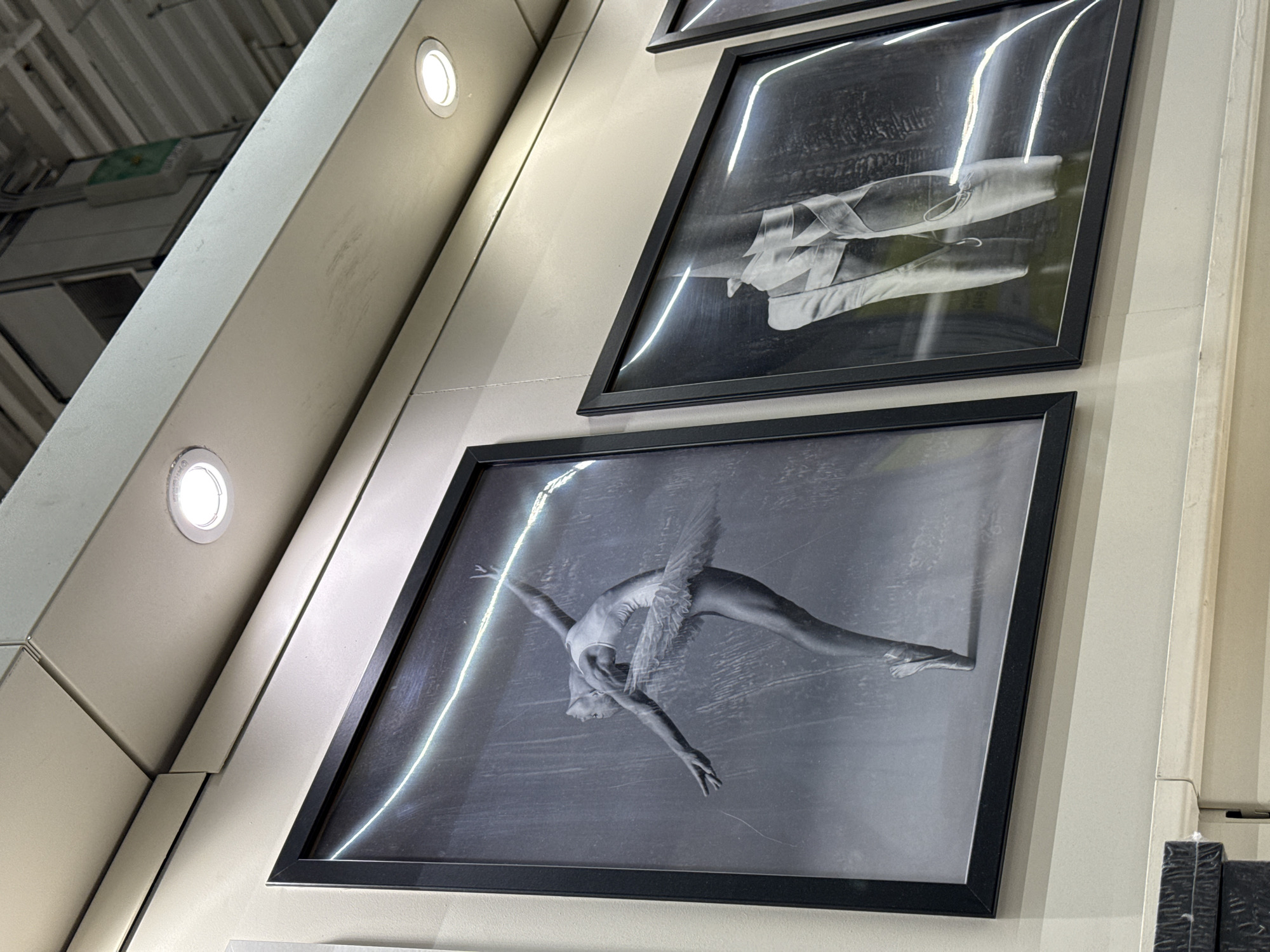 The image shows a wall in a building, possibly a gallery or theater, with two framed black and white photographs hanging on it. The top photograph depicts ballet shoes. The bottom photograph depicts a ballerina in motion. The building's architectural elements, including a light fixture, are visible on the left side of the frame. The environment suggests an indoor space with controlled lighting and a focus on artistic display.