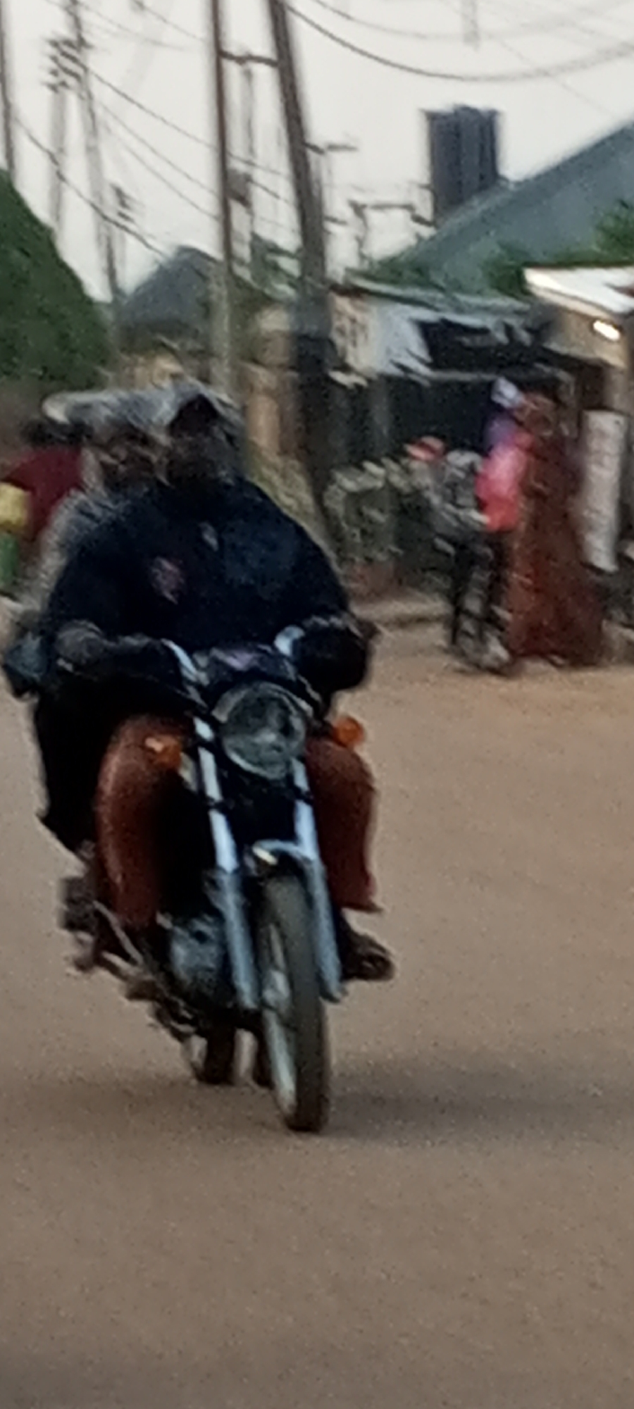 A man on a motorcycle is the primary subject in the image. He wears a dark garment and is positioned in the center of the frame. His face is obscured by shadow. The motorcycle, with a silver front and red panels, is moving forward. The street appears unpaved, with a light brown, sandy texture. In the background, there are indistinct buildings and people; utility poles and power lines are visible against the sky. The lighting is subdued, suggesting either early morning or late afternoon. This scene is set in Sabon Tasha, Nigeria.