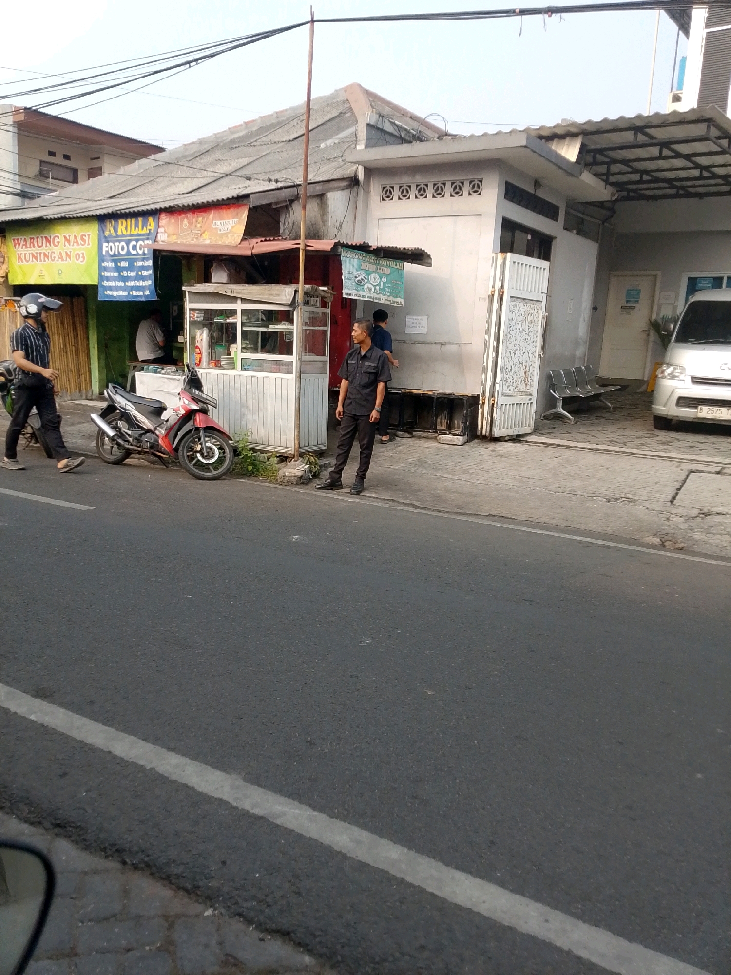 On a paved street in East Jakarta, Indonesia, a motorcycle is parked by a small shop. The shop has signs that read "WARUNG NASI KUNINGAN 03" and "R RILLA FOTO COPY". A man in a black shirt and pants stands to the right of the shop, and another man in a striped shirt and black pants walks past the motorcycle. The street is lined with buildings and utility poles.