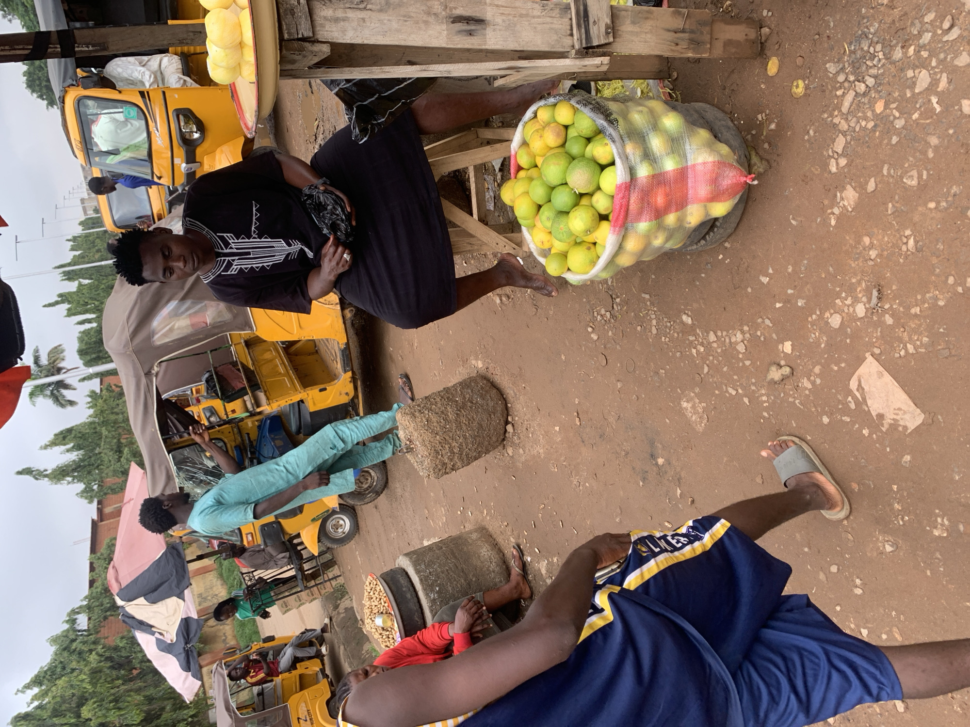 The image depicts a street market scene in Abuja, Nigeria, characterized by a dusty, reddish-brown ground. In the foreground, a man wearing a dark patterned tunic and black skirt is seated on a wooden stool, holding a black plastic bag. In front of him, a large, clear plastic bag filled with lemons and limes rests on a tire. To the left, another person, partially visible, is wearing a blue and yellow athletic outfit. In the background, several yellow auto-rickshaws are parked, some with passengers. One auto-rickshaw is partially covered by a large, multi-colored umbrella. A woman in a red top is seated next to a pile of peanuts on a large, round object. The sky is overcast.