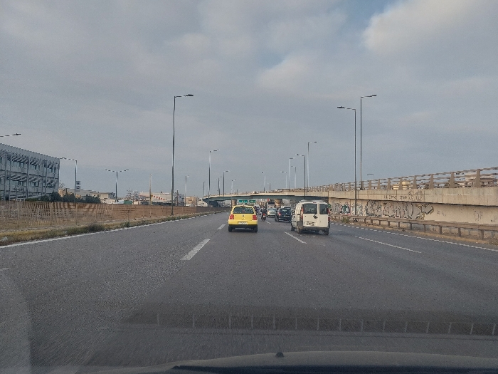 The image presents a daytime view of a multi-lane highway in Egaleo, Greece, under an overcast sky, observed from within a vehicle looking forward. The asphalt road features white dashed lane markings, indicating multiple lanes of travel in the same direction.
On the left side of the highway, a dry, unkempt strip of ground is separated from the road by a low, light-colored barrier composed of vertical slats. Beyond this, a modern, multi-story building with a facade incorporating horizontal reflective elements or louvers is prominent. Further in the distance, other urban buildings are discernible.
On the right side of the highway, a large concrete overpass or bridge crosses overhead, supported by concrete columns. The underside of the bridge's retaining wall is covered in extensive, varied graffiti. A light-colored concrete railing system tops the bridge deck. A green road sign is partially visible beneath the bridge on the far right. Numerous tall, black streetlights with single luminaires line both sides of the highway, extending into the distance.
Traffic consists of several vehicles. In the right lane, a white commercial van is observed from the rear, with other cars following it. In the left lane, a yellow sedan, consistent with a taxi, is visible ahead of the white van, also seen from the rear. Behind the yellow sedan, a motorcycle and additional vehicles are present. All vehicles are proceeding in the same direction. No pedestrians are visible. The scene suggests moderate vehicular traffic on an urban thoroughfare.