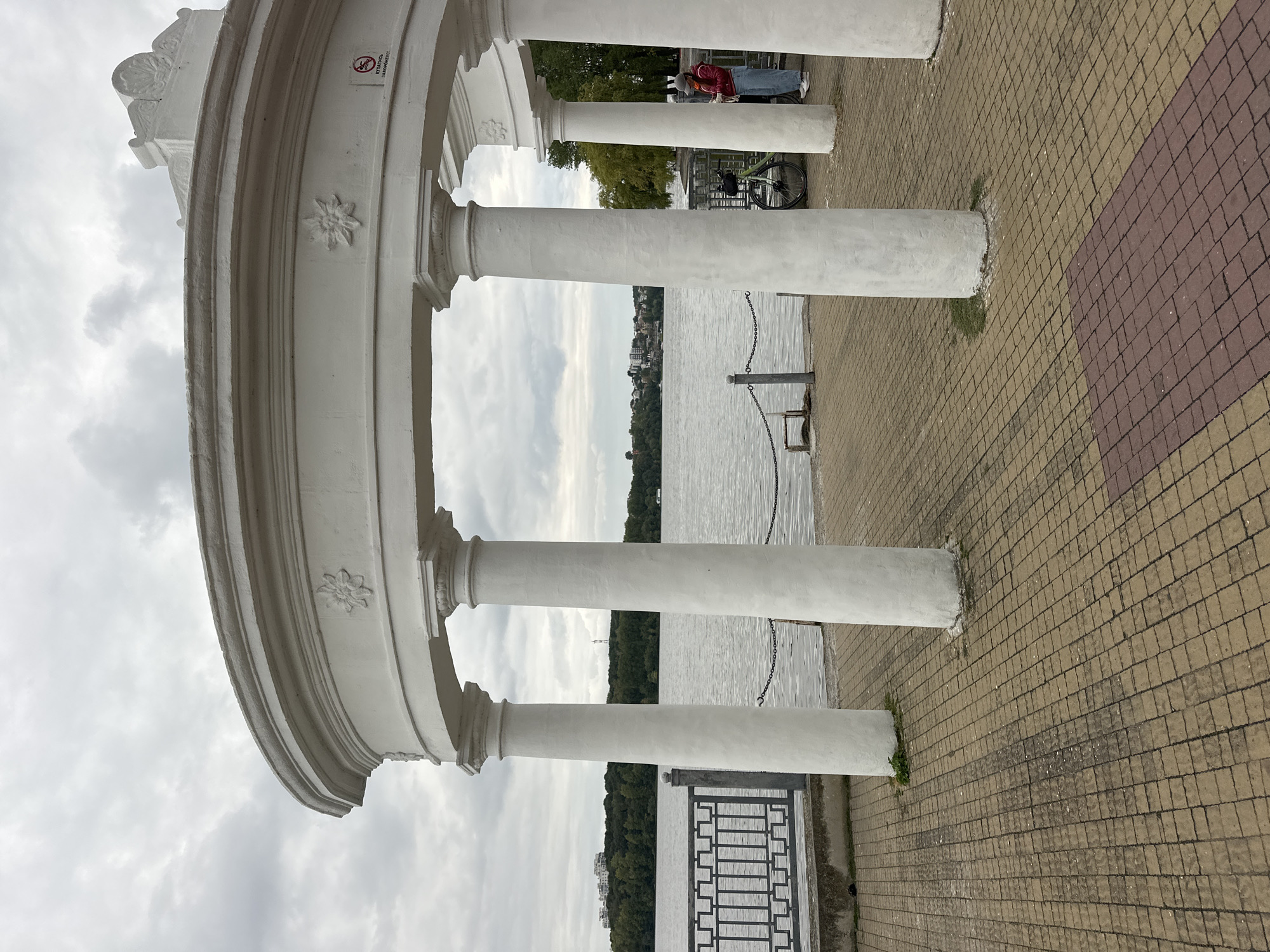 The image depicts a cloudy day at a waterfront promenade in Ternopil, Ukraine. In the foreground, a white, semi-circular rotunda with multiple columns stands on a paved walkway. The walkway is constructed with interlocking tan and maroon square tiles. The rotunda features decorative floral carvings on its archway and a "No Smoking" sign on its right side. Beyond the rotunda, a body of water, likely Lake Ternopil, stretches out, with a line of trees and buildings visible on the opposite shore. A metal fence separates the walkway from the water. In the middle ground, a bicycle is parked next to one of the rotunda's columns, and a person wearing a red jacket, blue jeans, and a cap is standing near the fence, looking out at the water. The overall scene suggests a public recreational area.
