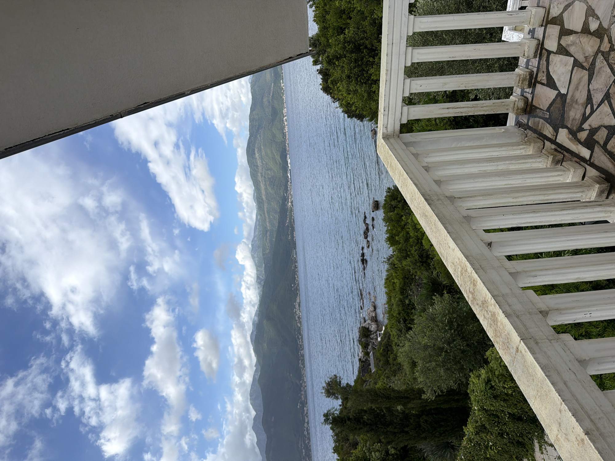 A view from a balcony in Gošići, Montenegro, features a body of water surrounded by mountainous terrain. The sky is blue with scattered clouds. A white balustrade runs along the balcony edge, and foliage is visible below. The balcony floor is paved with stone tiles. The water shows some ripples, and the mountains are lush with greenery.