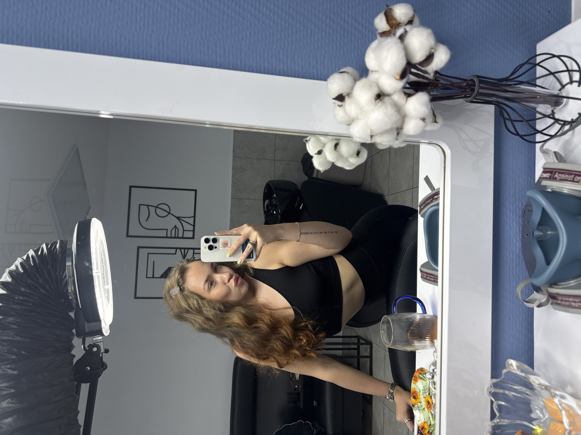 An indoor mirror selfie captures a young woman posing in what appears to be a dressing room or vanity area. The woman, with long, wavy brown hair styled with clips, is seen from the chest up, wearing a black sports bra or crop top and black leggings. She holds a white iPhone with multiple cameras to take the picture, showcasing light-colored nail polish, rings, a bracelet, and a watch. Her expression features slightly pursed lips and a direct gaze at the phone camera. Small, illegible text is tattooed on her right forearm.
The room reflected in the large white-framed mirror features white walls adorned with two black-framed abstract line art pictures. A large ring light with a flexible black arm stands to the left. Dark furniture, including a chair and a table, is also visible in the reflection.
Outside the mirror's reflection, the background consists of a textured blue wall. On the white vanity surface next to the mirror, a bouquet of white cotton branches is arranged in a black wire whisk-like vase. Other items include a clear glass mug with a blue handle, a decorative plate with a sunflower pattern, and a blue object resembling goggles or a mask. Partially visible text on these items reads "own," "Against or," and "image." The setting suggests a well-lit, possibly professional or dedicated personal space.