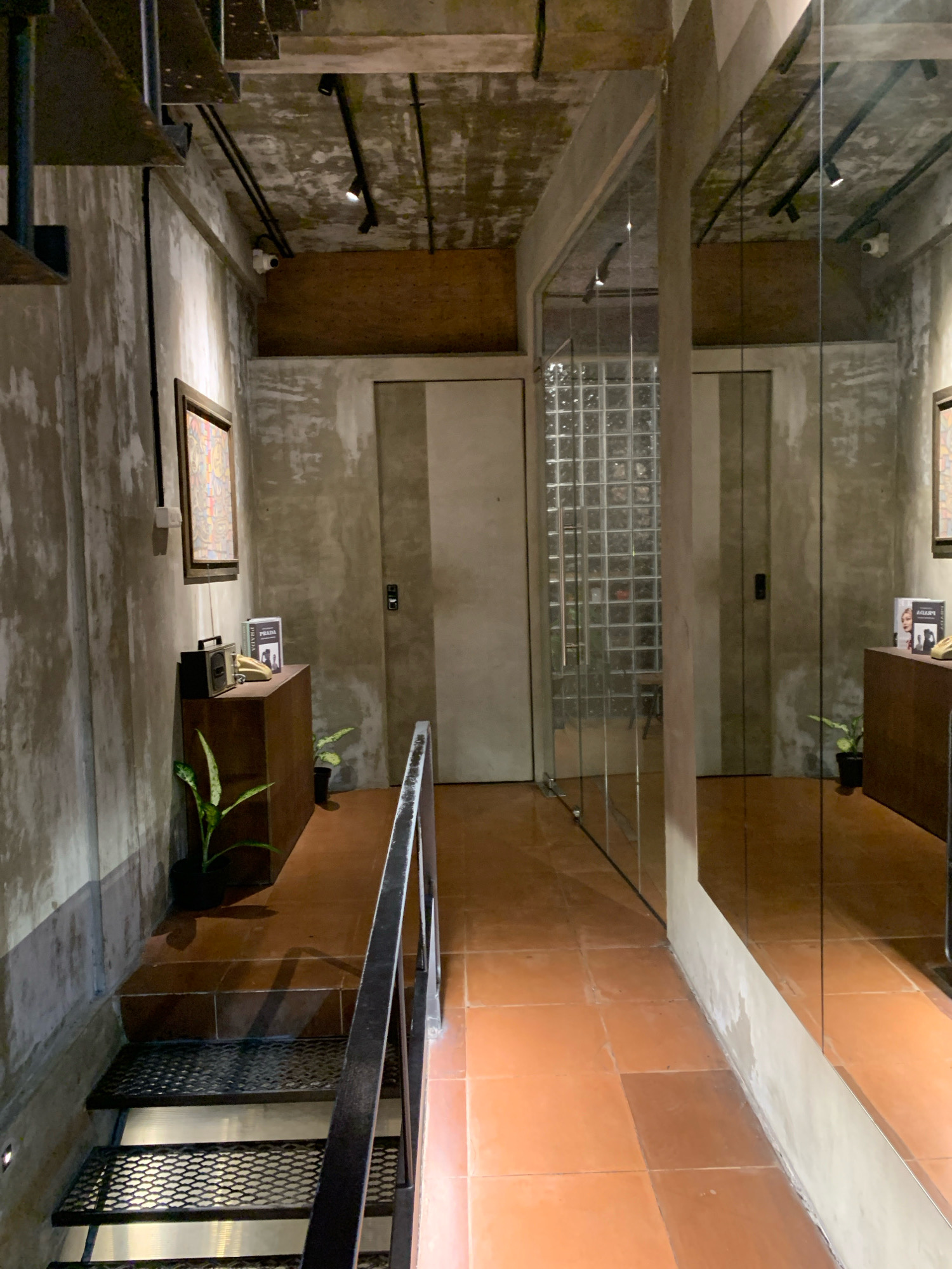 The image depicts an indoor space, likely a hallway or entrance, with a utilitarian and industrial aesthetic. The walls are unfinished concrete with visible imperfections, and the ceiling features exposed beams and conduits. Black track lighting illuminates the area. To the left, a staircase with black metal grating steps descends, bordered by a black metal handrail. A framed artwork hangs on the concrete wall above the staircase. Below the artwork, a dark wooden console stands, topped with a vintage radio, a yellow rotary phone, and two books. Two potted plants, one on the console and another on the floor, add a touch of greenery. Further down the hallway, to the right, a large mirror reflects the opposite wall, which also appears to be concrete and features a glass block partition. Two doors are visible, one with a push plate and handle, and another that is closed and flush with the wall. The flooring consists of terracotta-colored square tiles. This scene is consistent with the interior design of a modern establishment, potentially a cafe, restaurant, or boutique, in the city of Mojokerto, Indonesia.